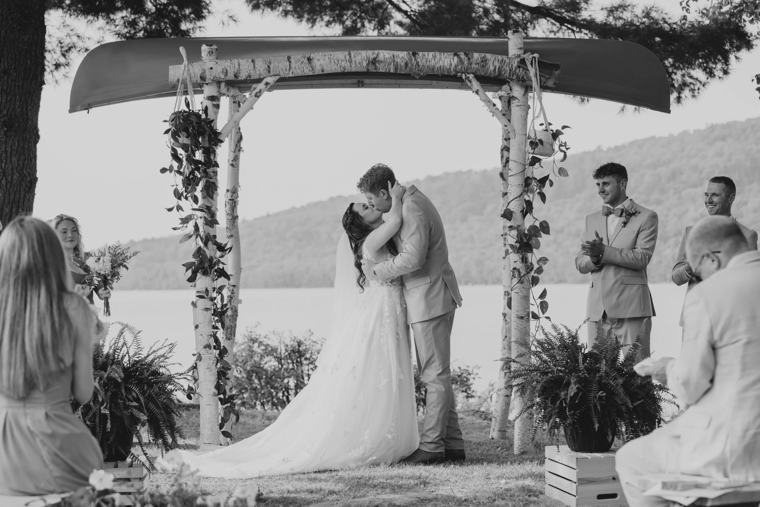 A bride and groom kissing under a rustic wedding arch outdoors, surrounded by guests. The scene is in black and white with trees and hills in the background.