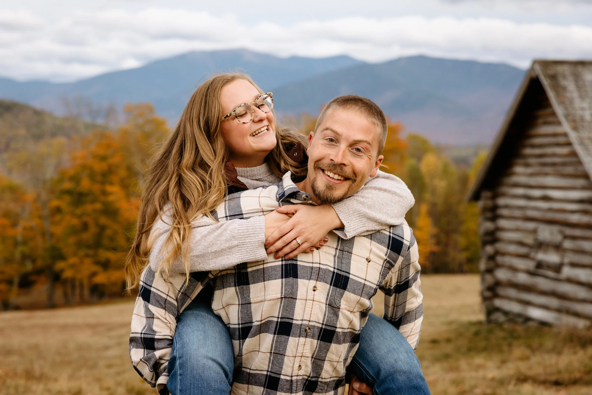 A smiling man giving a piggyback ride to a woman in a rural outdoor setting with autumn trees and mountains in the background.