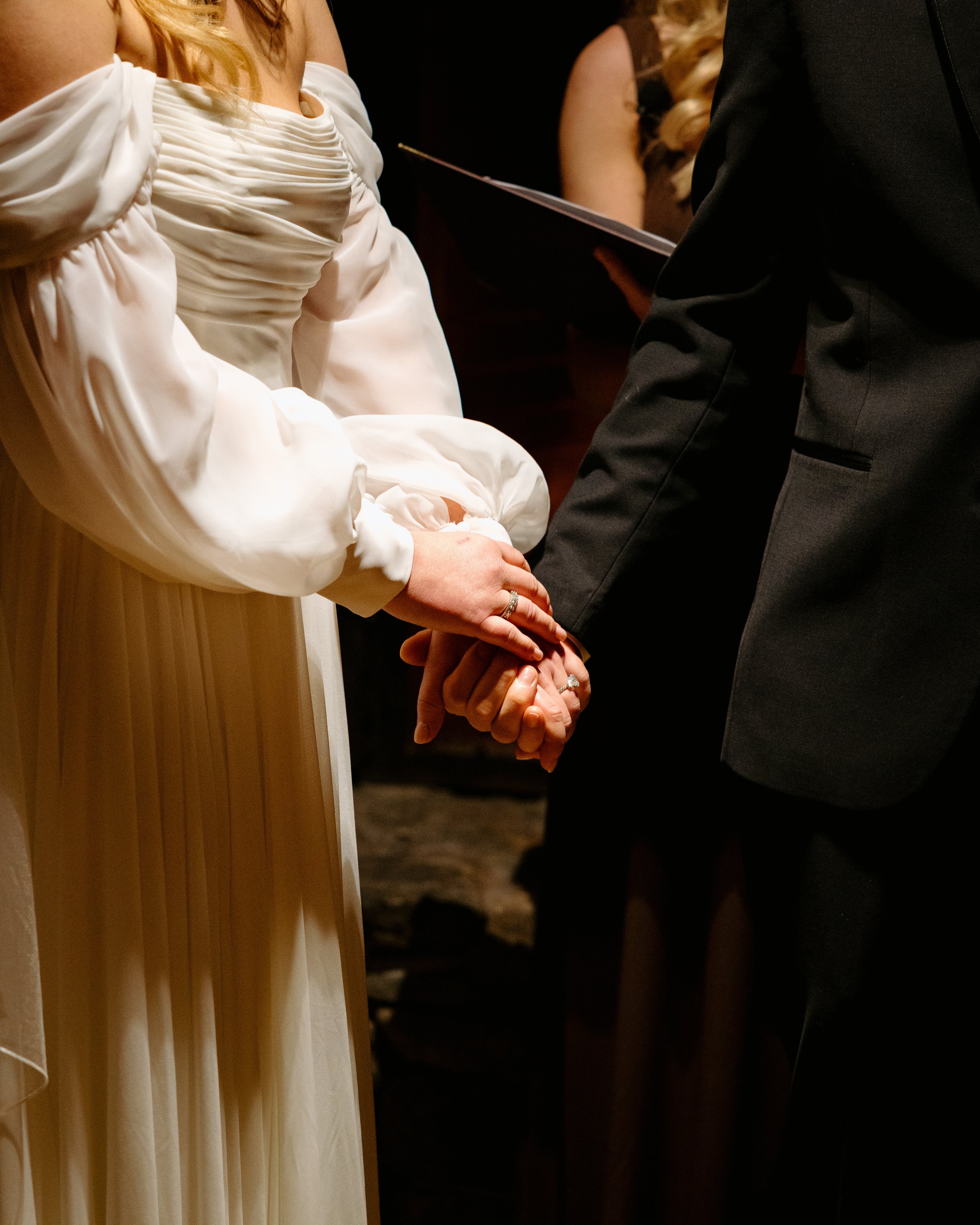 A close-up scene of a wedding ceremony focusing on a bride and groom holding hands, with the bride wearing a white dress with ruched detailing at the neckline and puffed sleeves, and the groom in a black suit. A person in a black dress is visible in 