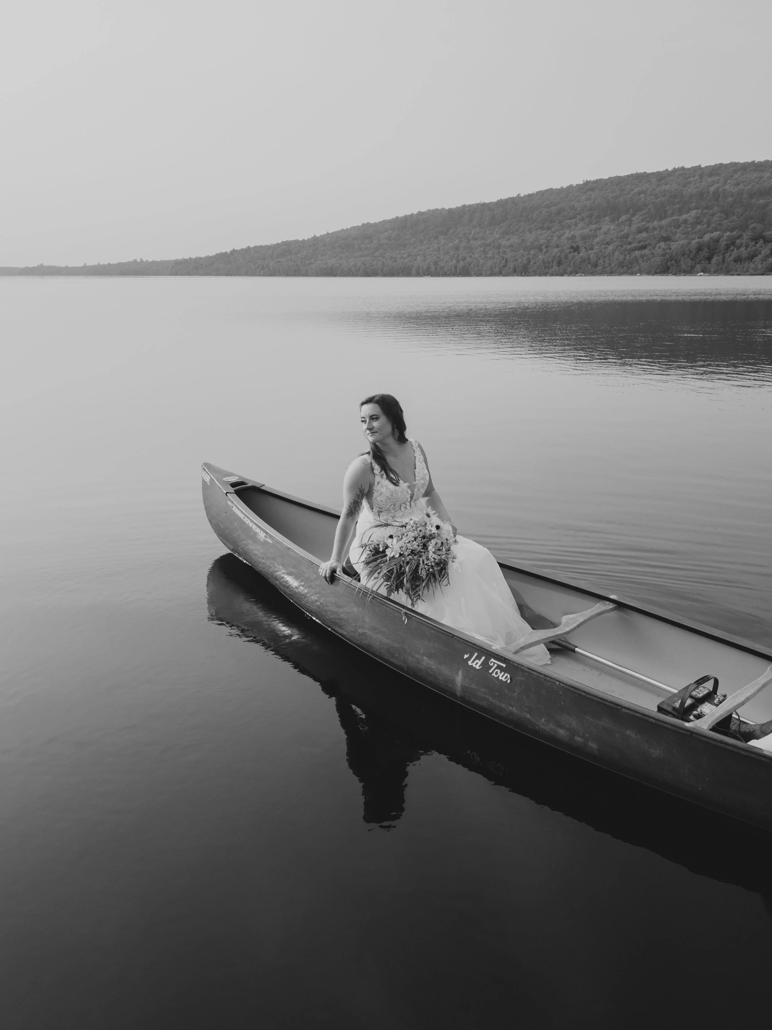 A woman in a wedding dress sitting in a canoe on a calm lake with a hilly landscape in the background, holding a large bouquet of flowers.