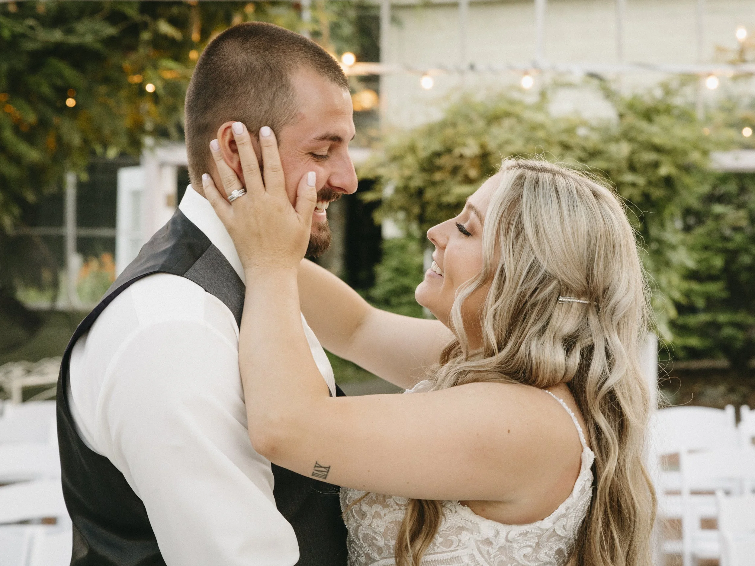 A couple dancing close together outdoors during a wedding, with the woman wearing a lace wedding dress and the man in a suit, both smiling lovingly at each other.