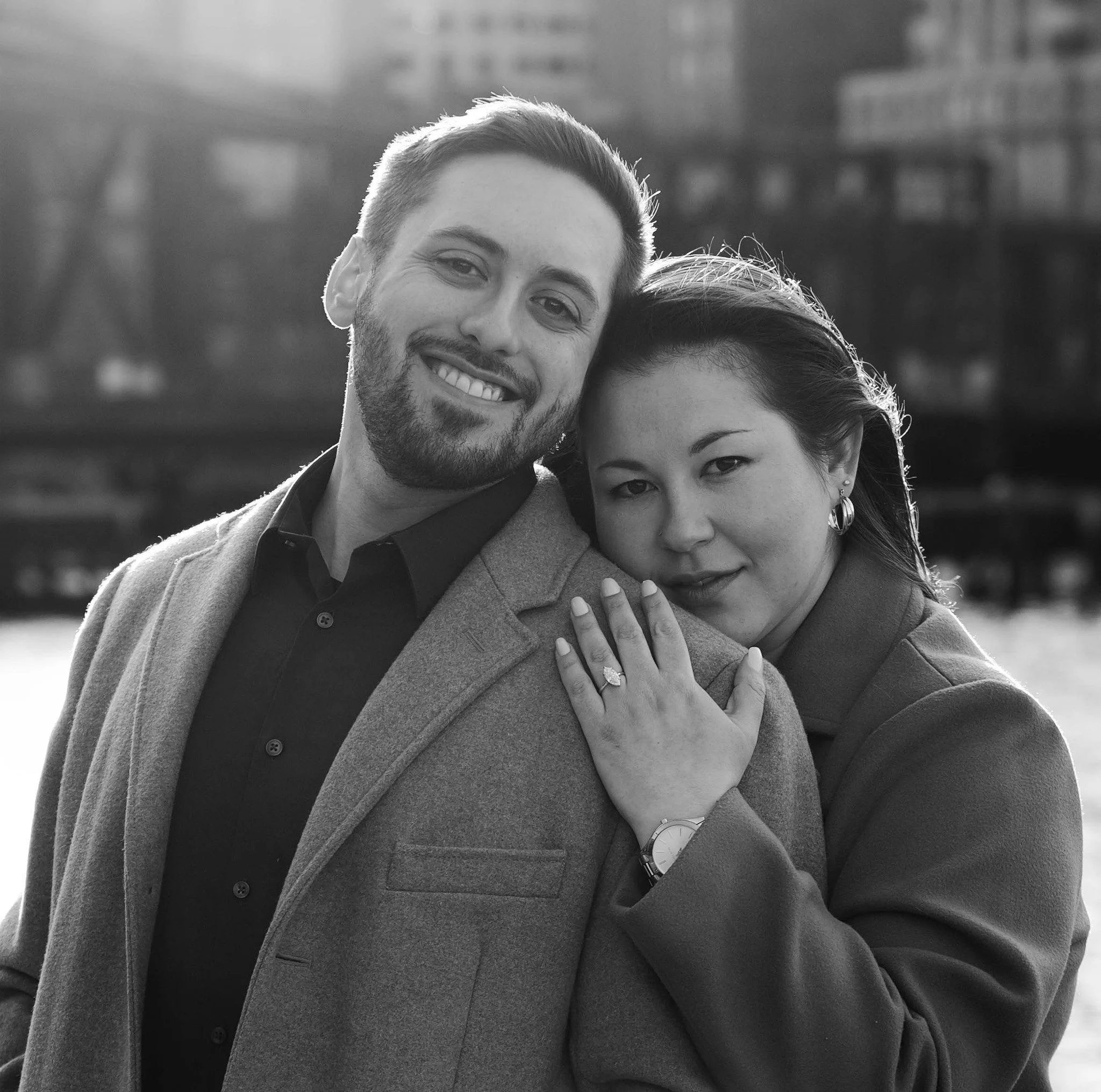 Close-up black and white photo of a smiling man and woman, with the woman showing her wedding ring. The man is wearing a watch, and both are wearing coats, standing outdoors with a blurred cityscape in the background.