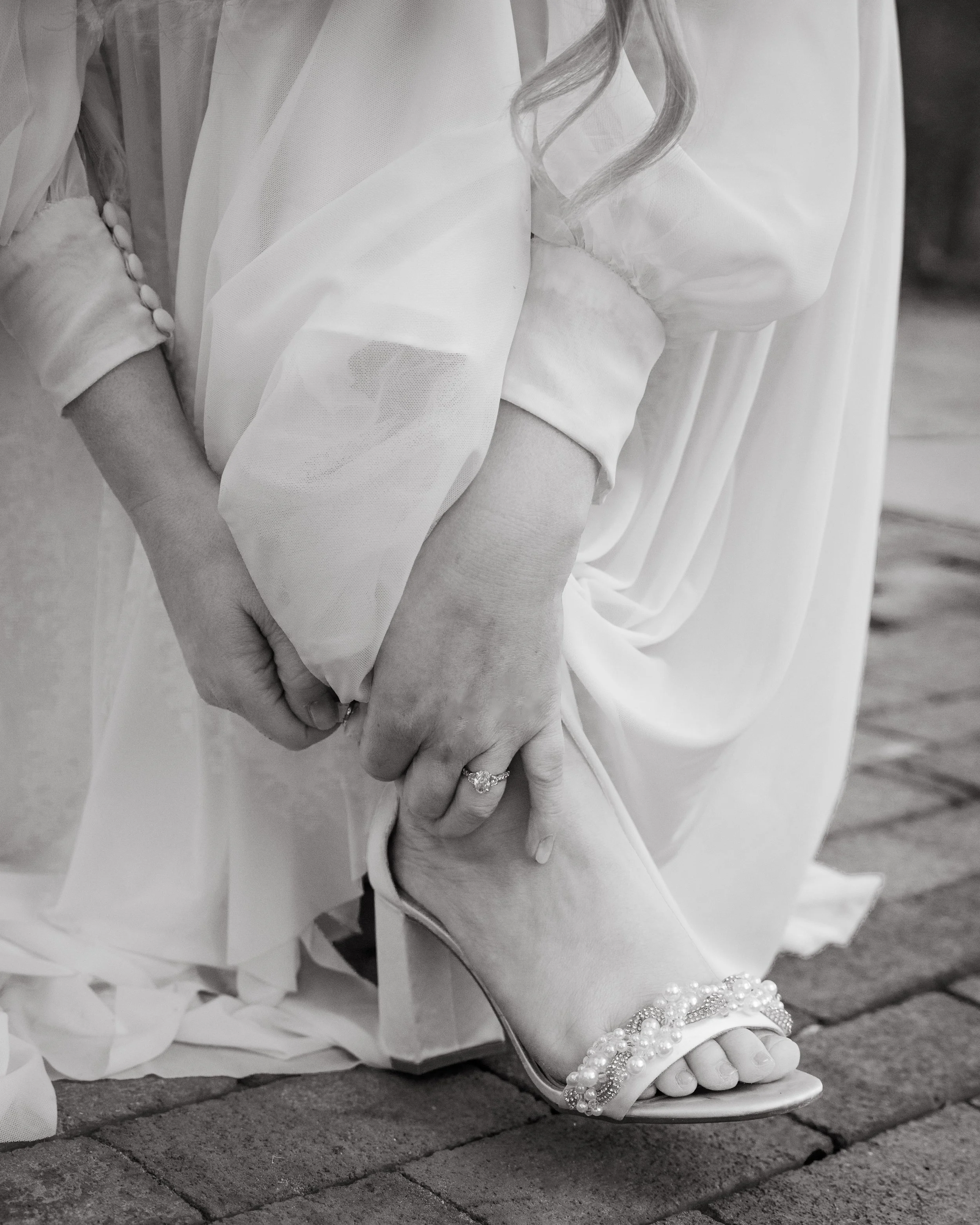 Close-up of a person wearing a white dress and jewelry, holding their foot with their hand. The focus is on the foot adorned with pearl jewelry and the back of the dress with buttons.