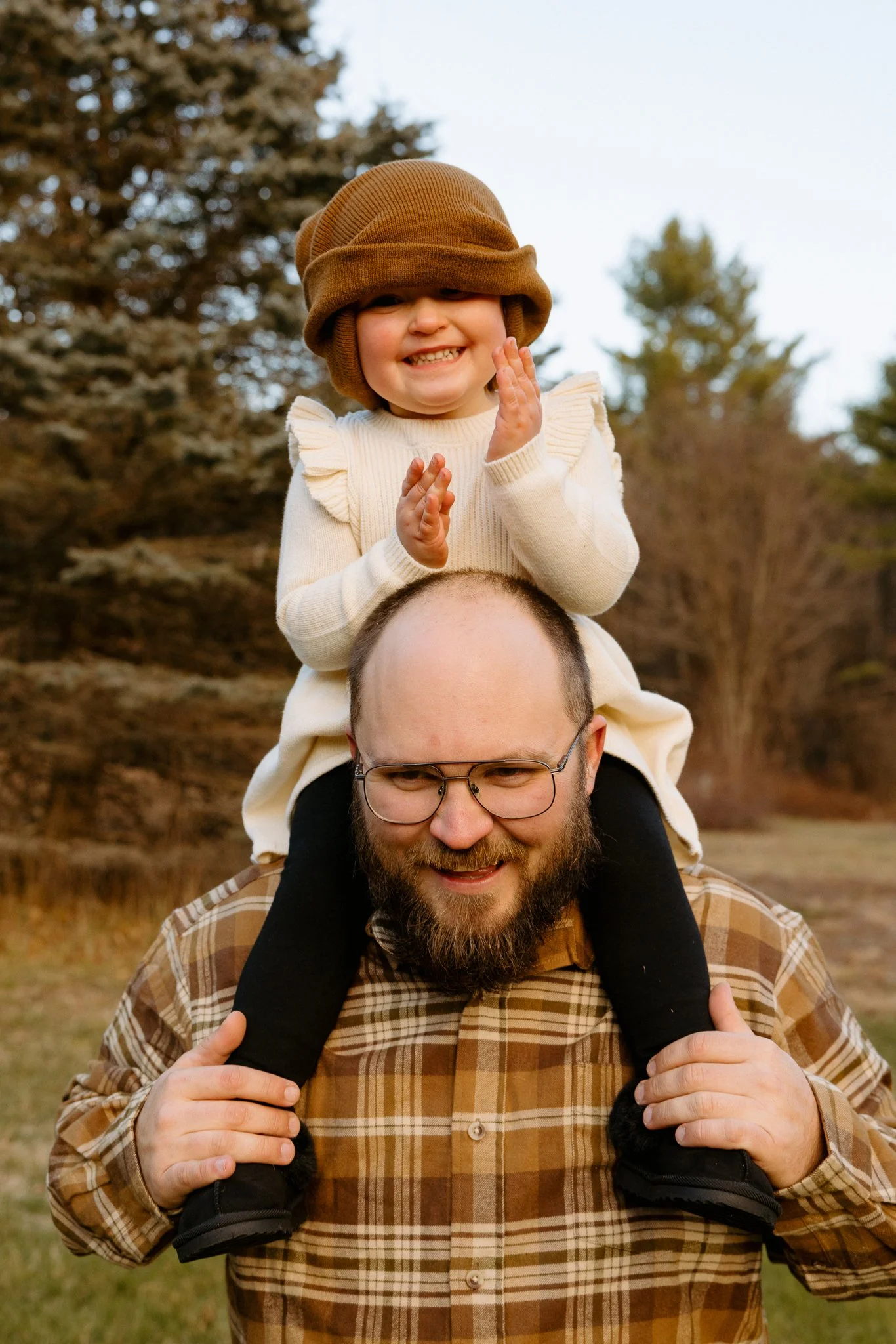 A man wearing glasses and a plaid shirt is carrying a young girl on his shoulders in a park with trees in the background. The girl is smiling and clapping, wearing a brown hat and a cream sweater.