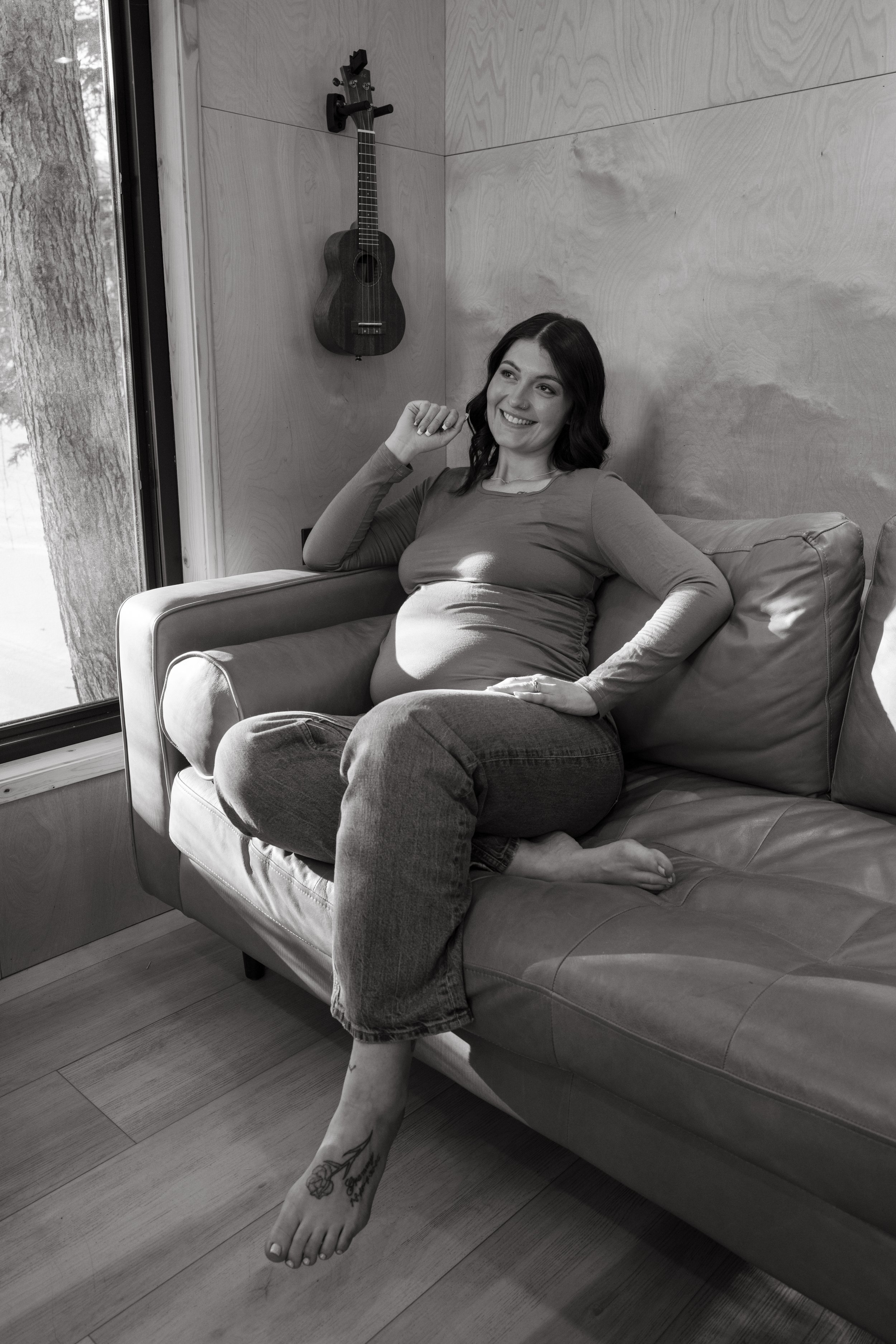 A smiling woman with dark, wavy hair sitting on a couch in a room with light wooden walls. A small ukulele hangs on the wall behind her. She is barefoot, wearing jeans and a long-sleeved shirt, with tattoos visible on her left foot and right arm.