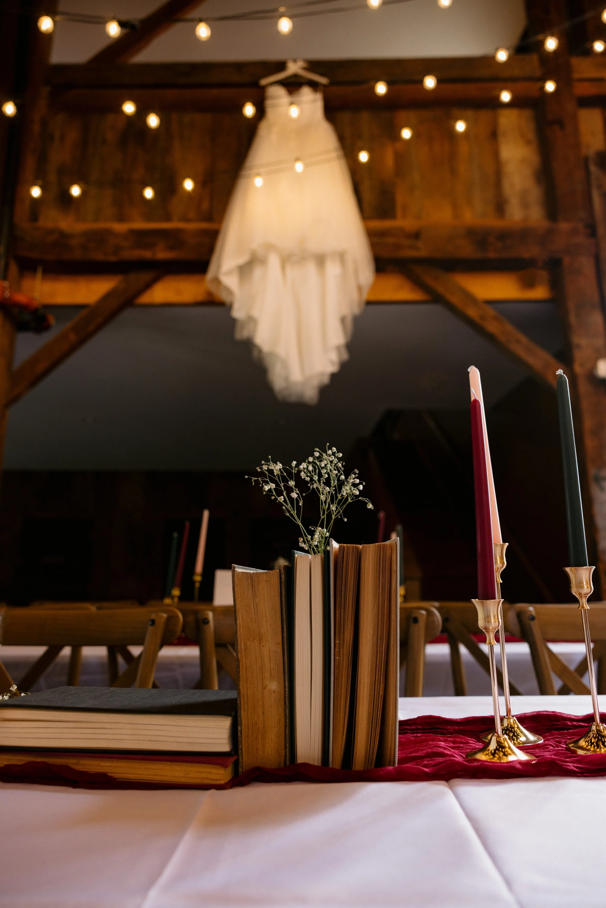 Close-up view of a table setting with books, a small vase with baby's breath flowers, and three tall candlesticks with red, white, and black candles, in a rustic wooden hall decorated with string lights and a hanging fabric lampshade.