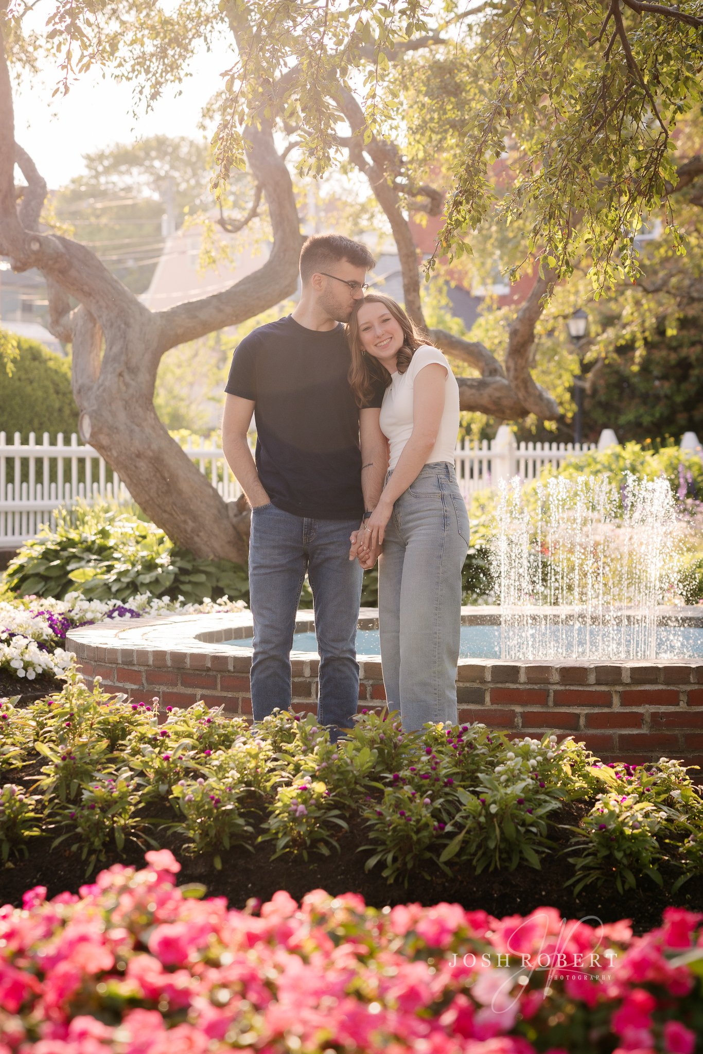 A smiling couple standing close together in a garden with colorful flowers and a fountain, surrounded by trees and a white picket fence, during sunset.