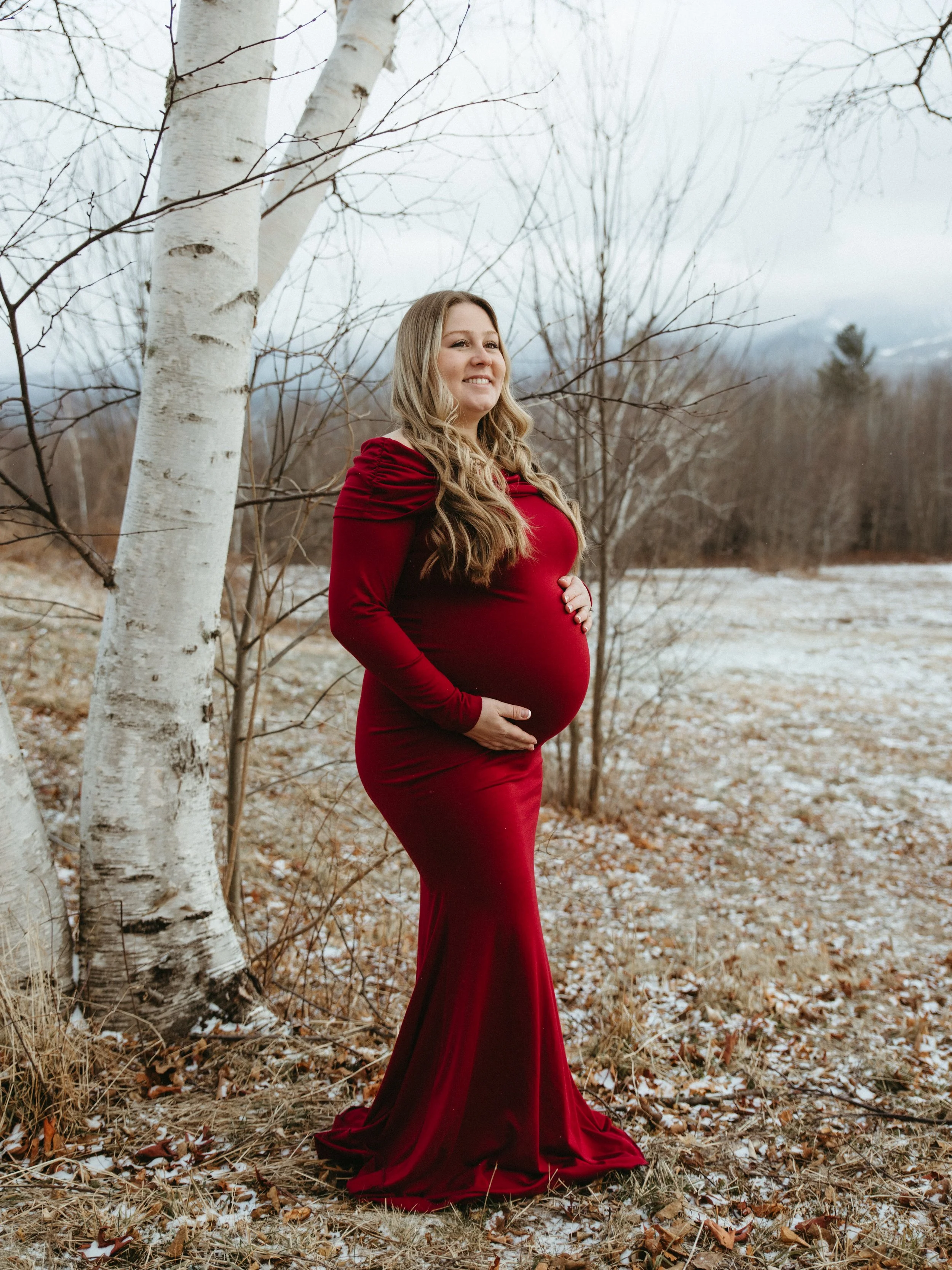 Pregnant woman in a red velvet gown standing outdoors in a winter landscape with bare trees and a light snow-covered ground.
