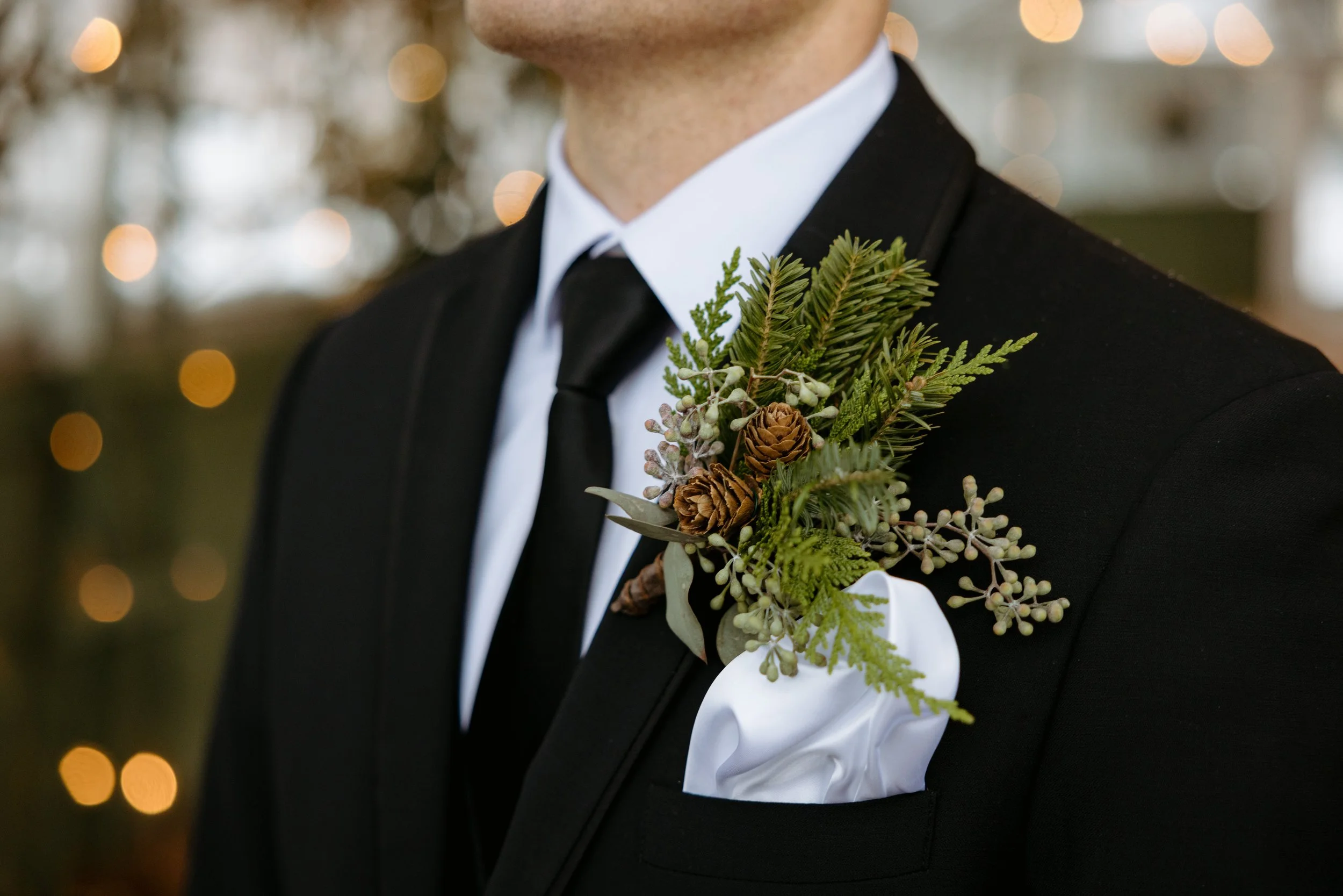 Close-up of a man dressed in a black suit with a white shirt and black tie, wearing a boutonniere made of green foliage, pinecones, and small berries, with blurred festive lights in the background.