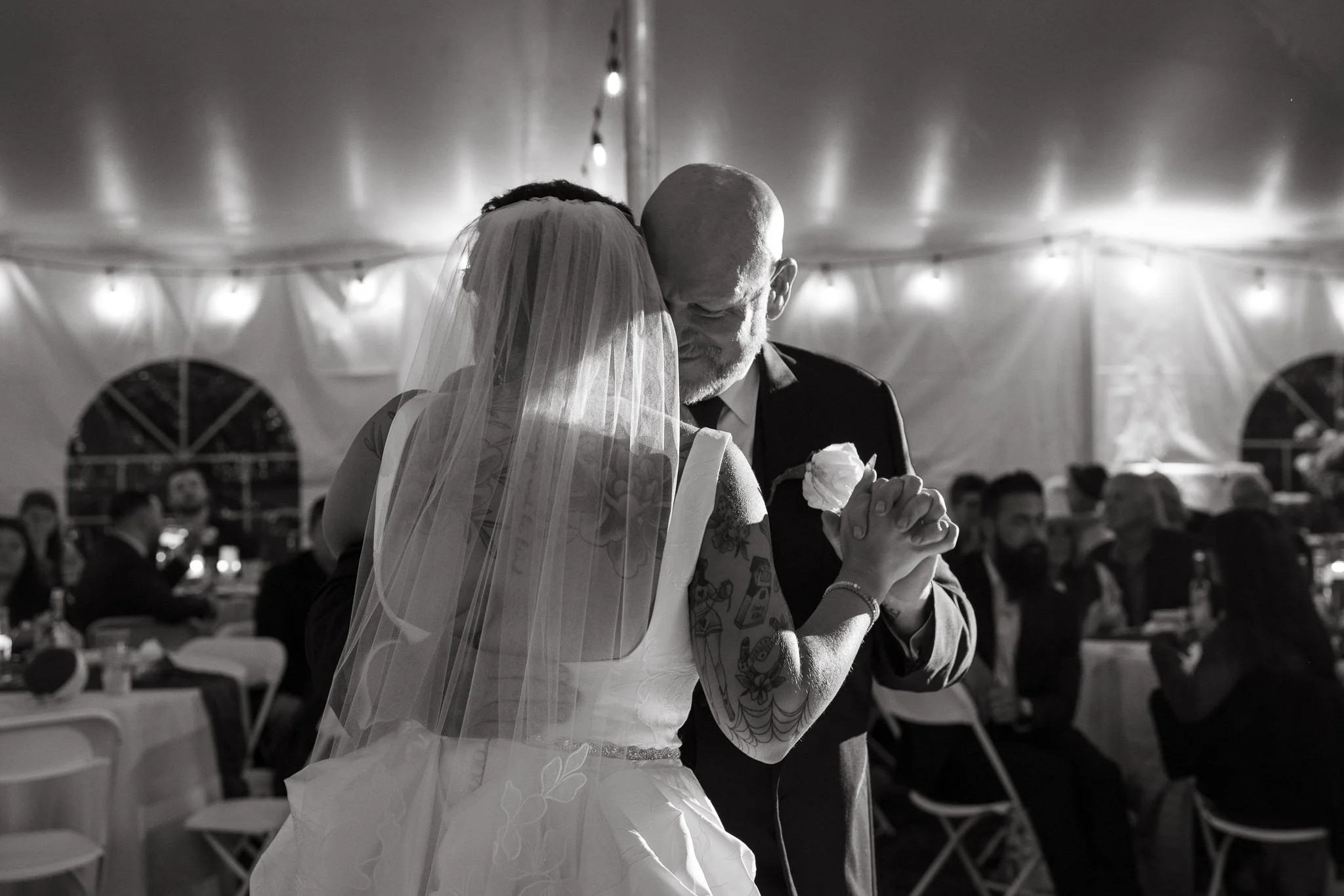 A bride and groom share a dance during their wedding reception inside a large tent, with guests seated at tables around them.