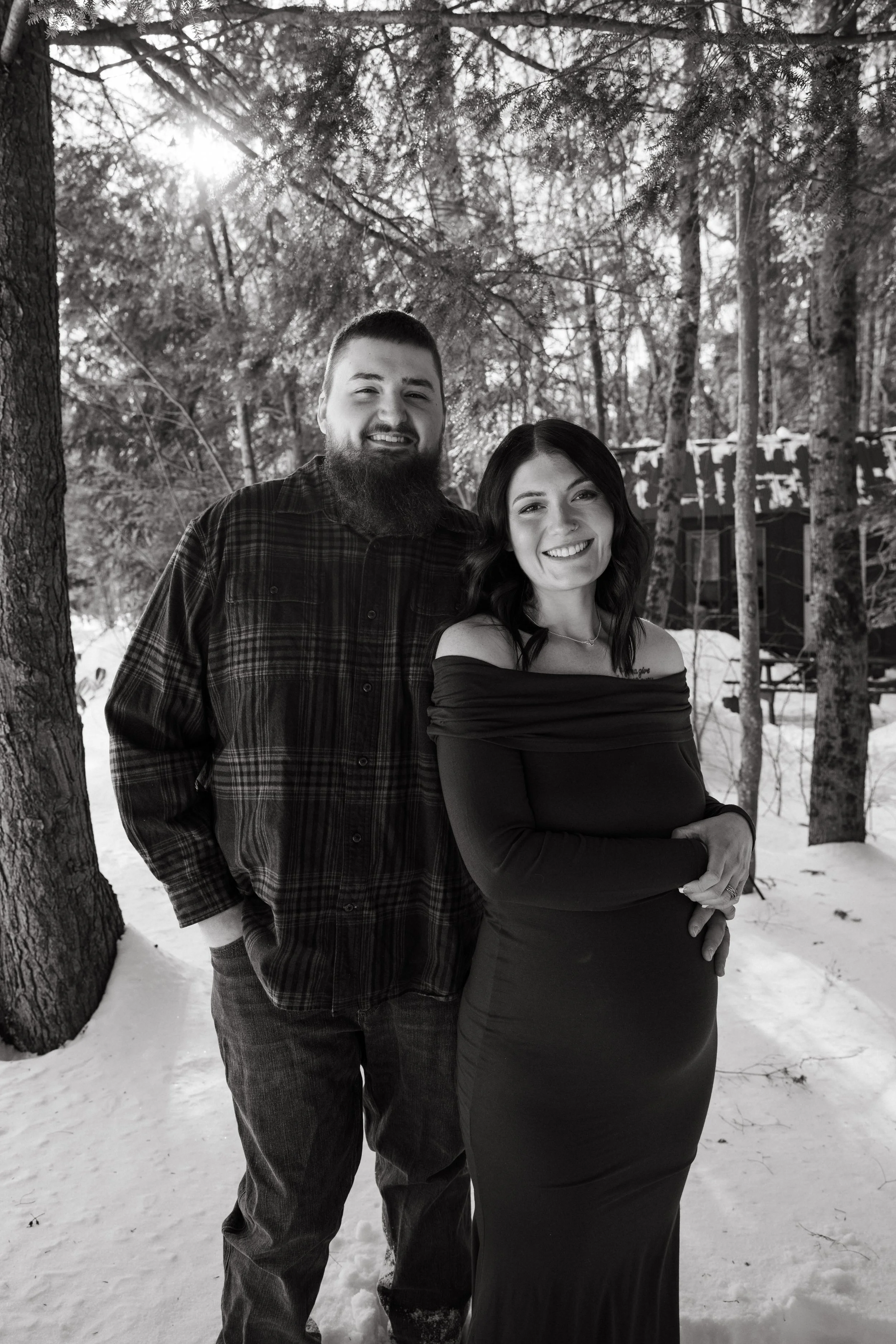 A black and white photo of a smiling man with a beard and a woman with dark hair, both standing outdoors in a snow-covered area with trees in the background. The man is wearing a plaid shirt, and the woman is wearing an off-the-shoulder dress.