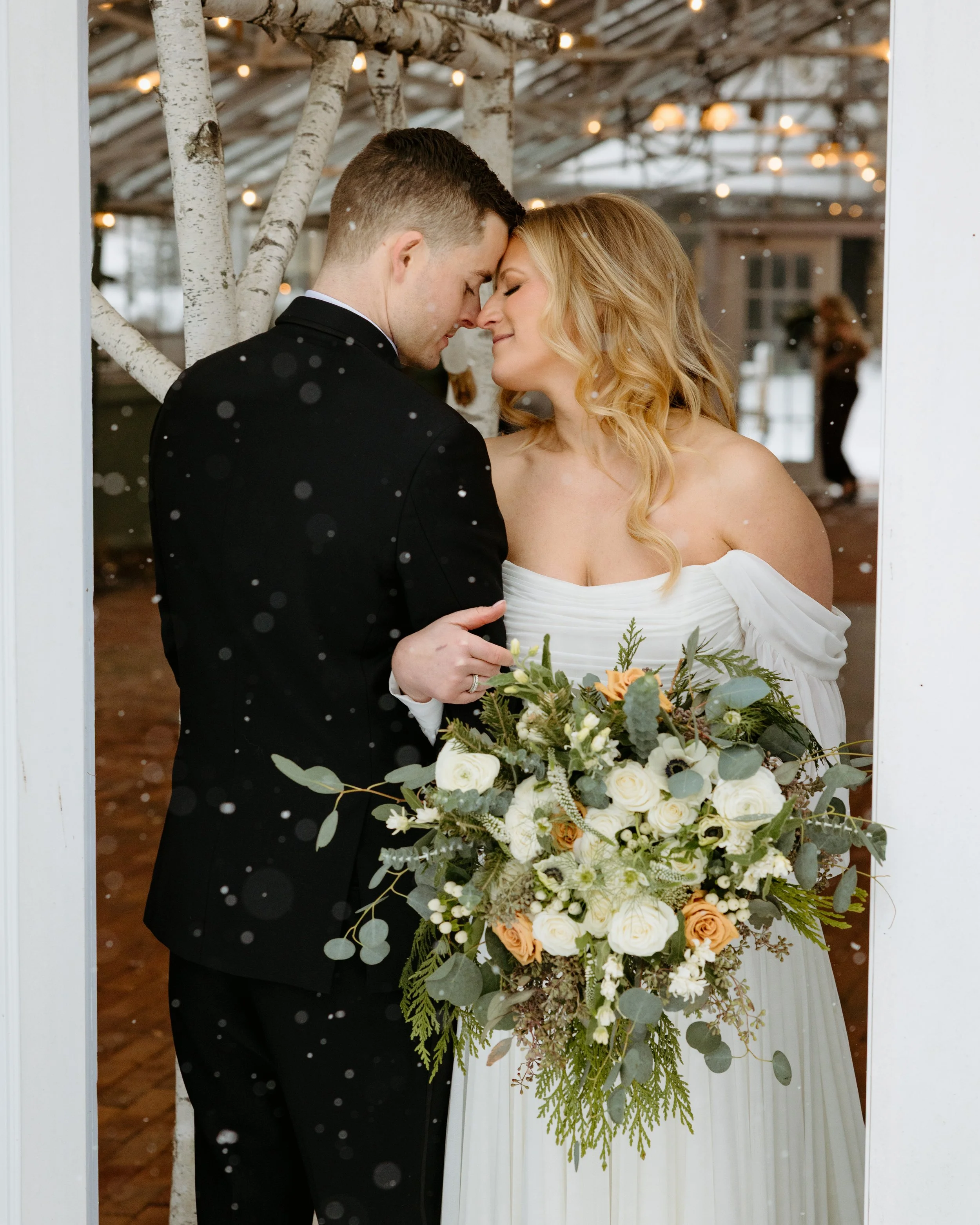 A bride and groom sharing a tender moment with foreheads touching inside a decorated venue, with the bride holding a large bouquet of white and cream flowers and greenery.