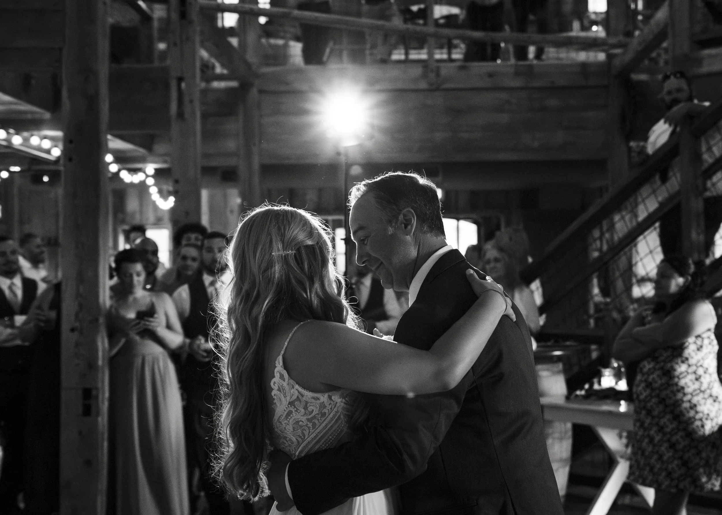 A black-and-white photograph of a dance couple sharing an intimate moment, with the woman wearing a lace dress and the man dressed in a suit, surrounded by other guests at a social event.