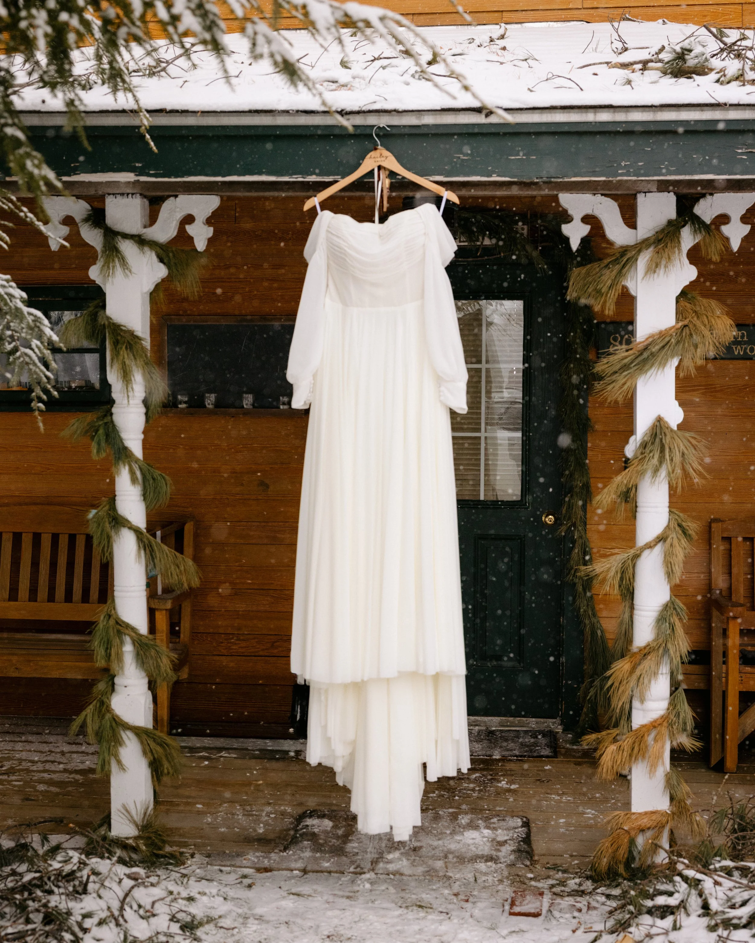 A white wedding dress hangs on a wooden hanger on a porch decorated with pine garlands and snow, with a wooden house exterior in the background.