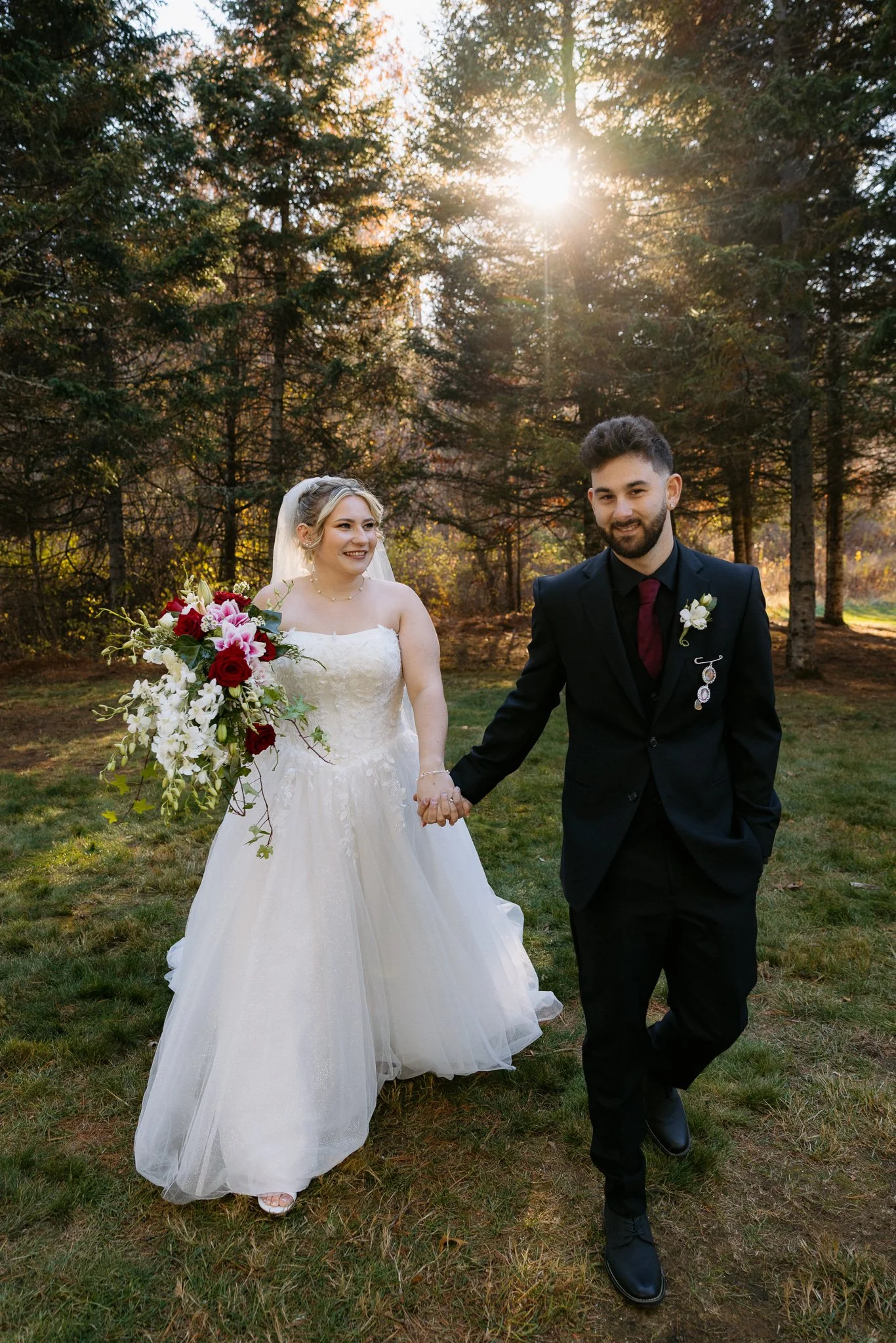 A bride and groom holding hands and walking outdoors in a forested area during sunset. The bride wears a white wedding gown and veil, carrying a bouquet of red, pink, and white flowers. The groom wears a black suit with a boutonniere and a burgundy tie. They are smiling and looking at each other.