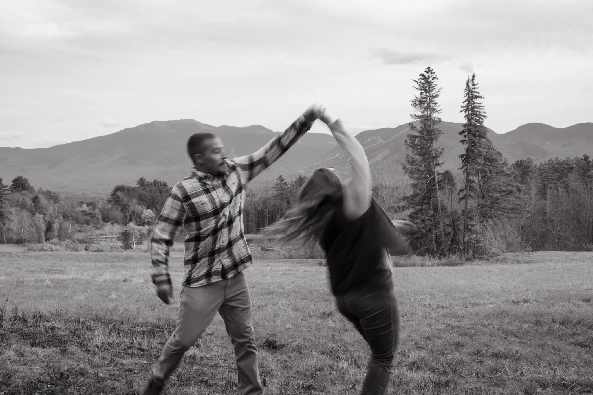 Two people, a man and a woman, dancing together in an open field with mountains and trees in the background, black and white photo.
