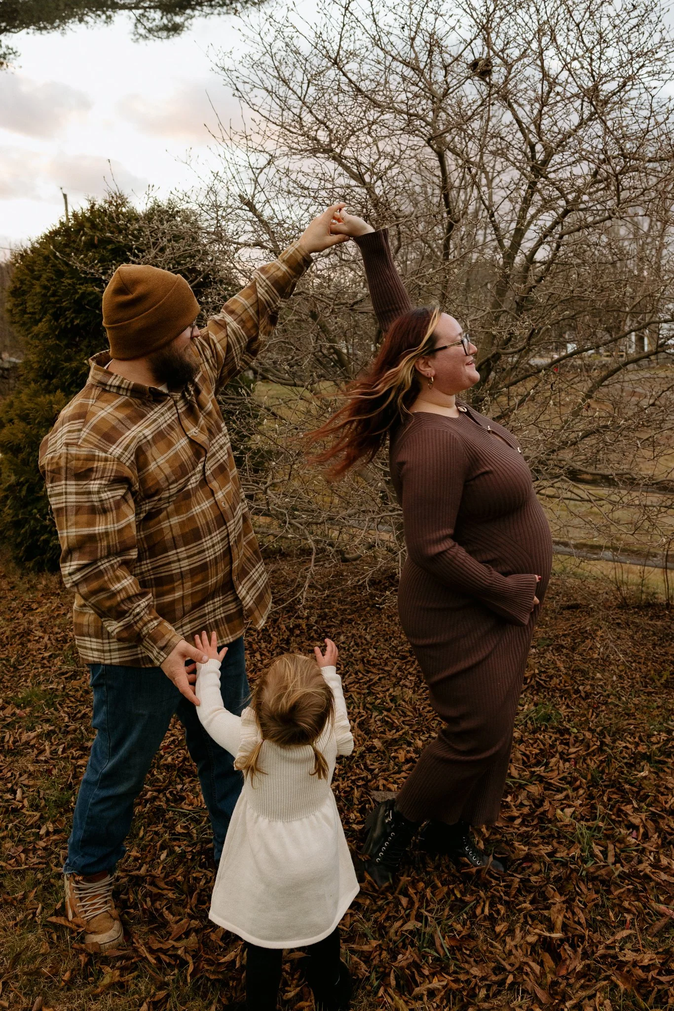 A group of three adults and two children outside on a fall day, with leafless trees and fallen leaves on the ground, holding hands and dancing together.