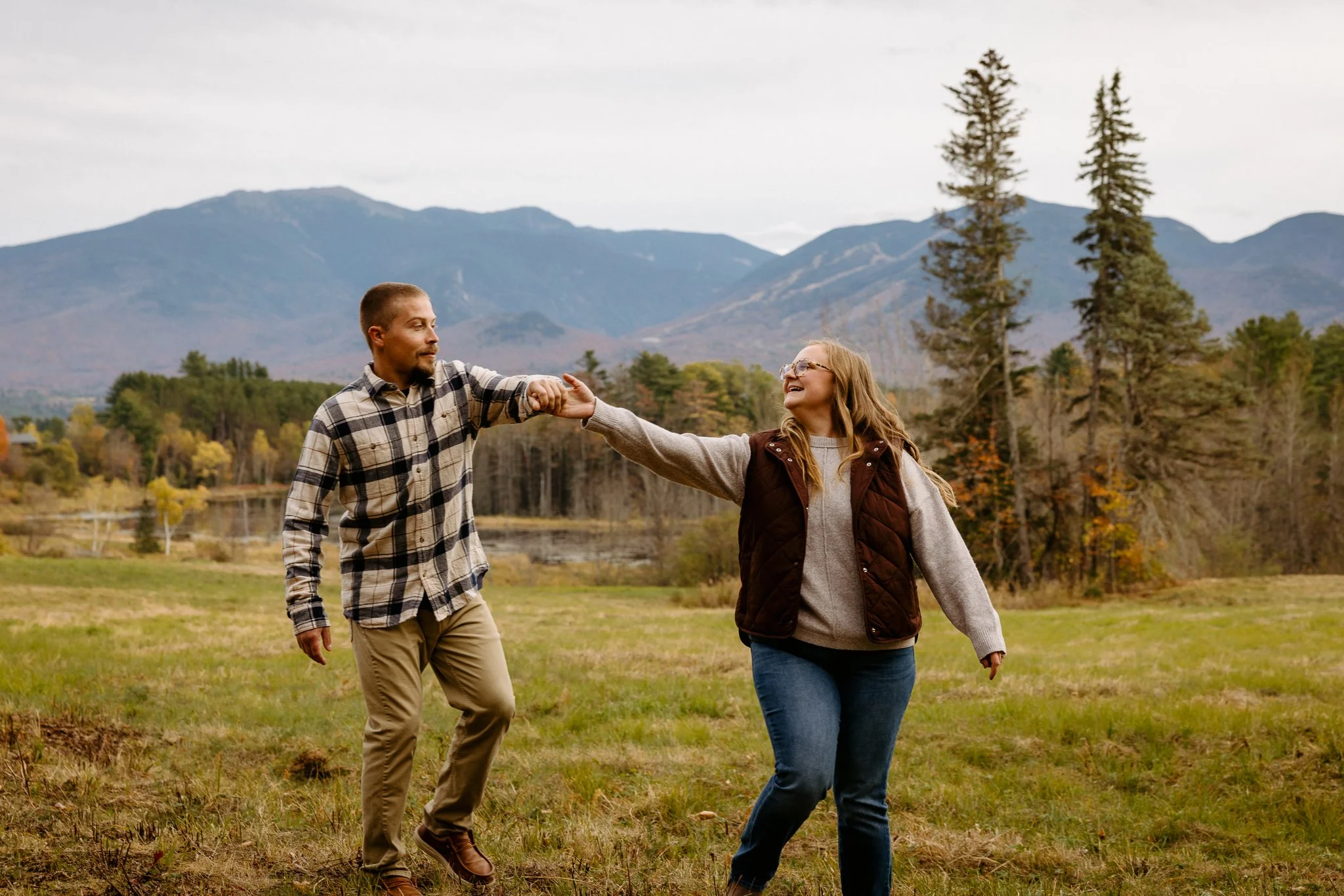 A man and woman walk outdoors during autumn, holding hands and smiling, with mountains, trees, and a cloudy sky in the background.