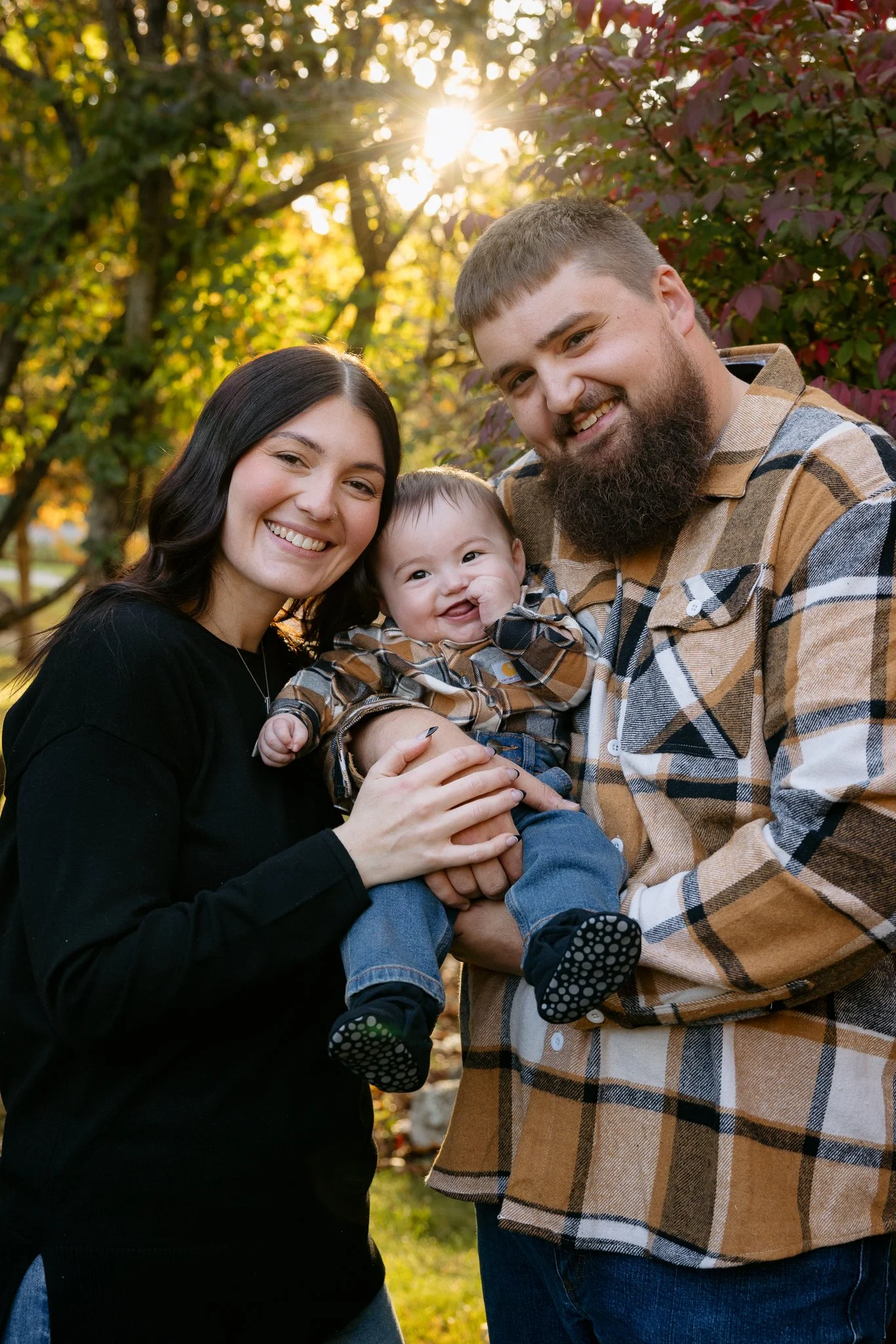 A family of three posing outdoors in a park during autumn, with trees and sunlight in the background. The parents are smiling, holding their young child between them.