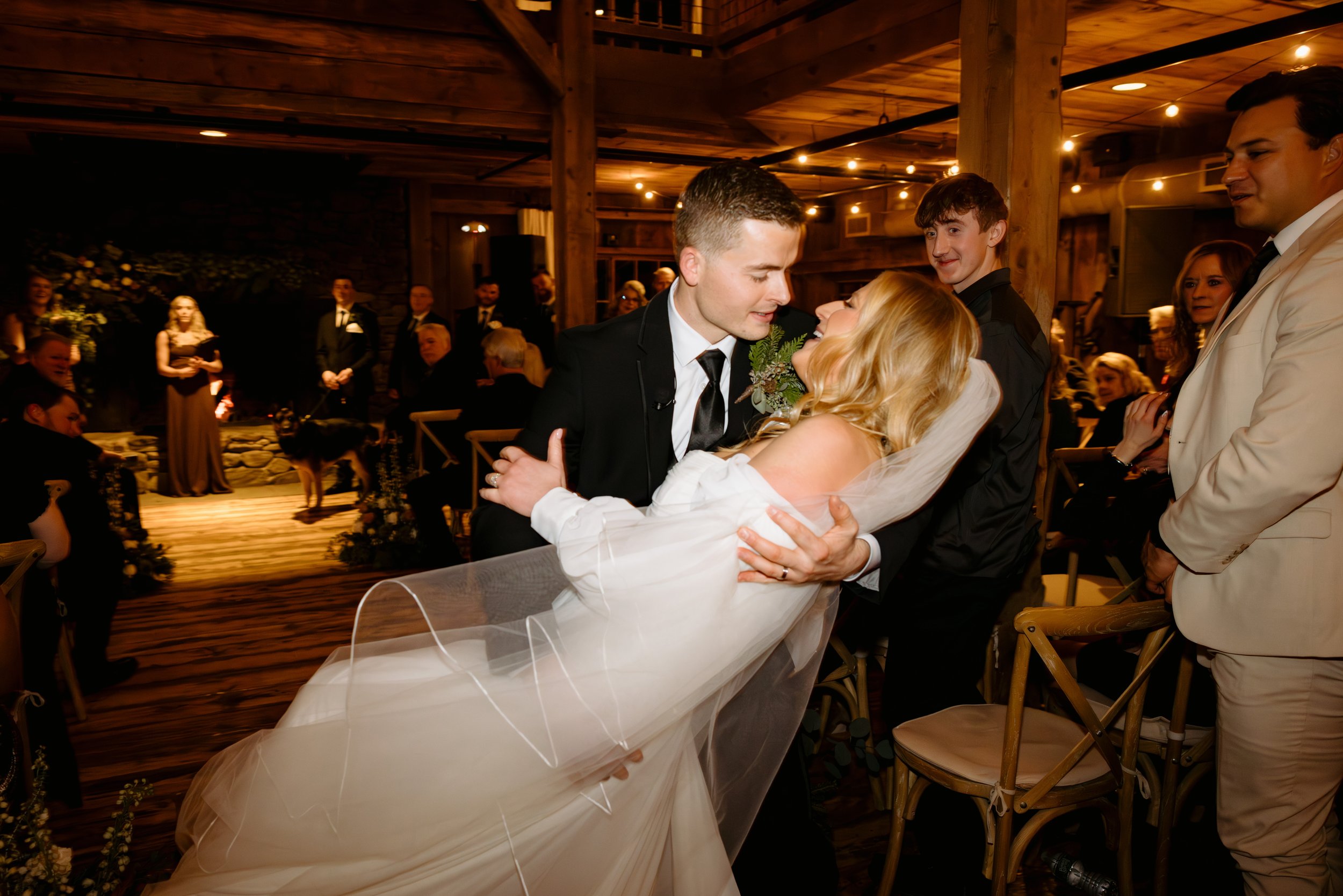 A bride and groom dancing at their wedding reception with friends and family watching in a rustic wooden venue decorated with string lights.