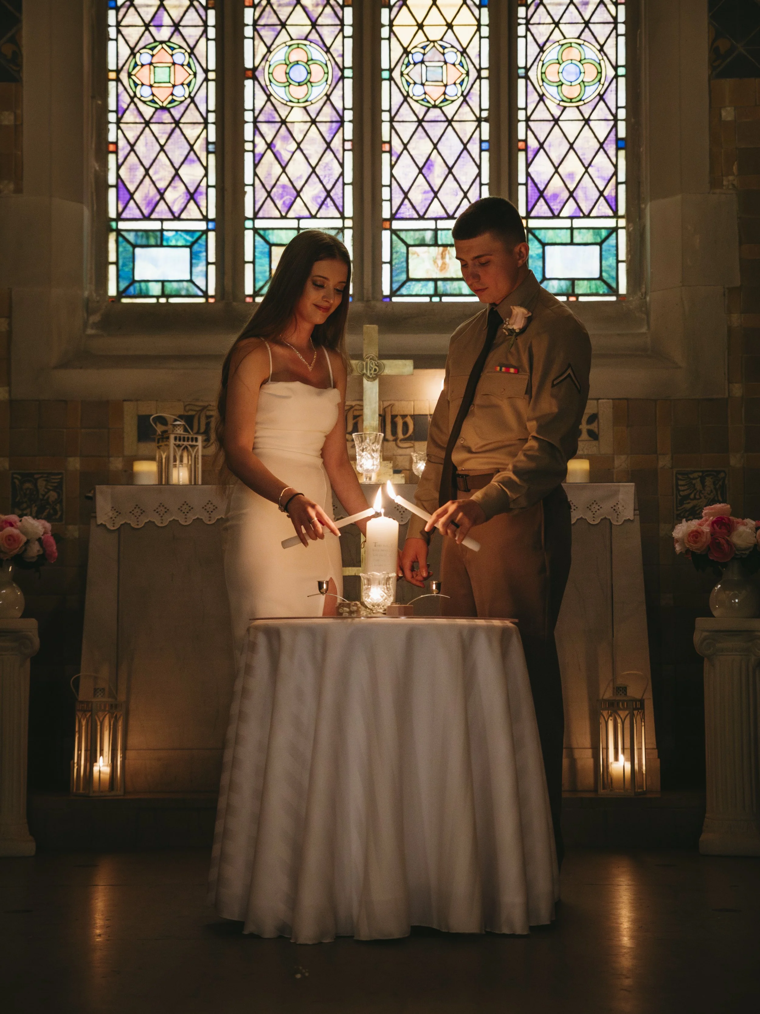 A young woman in a white dress and a young man in a military uniform stand inside a church, lighting candles during a ceremony with stained glass windows and a cross in the background.