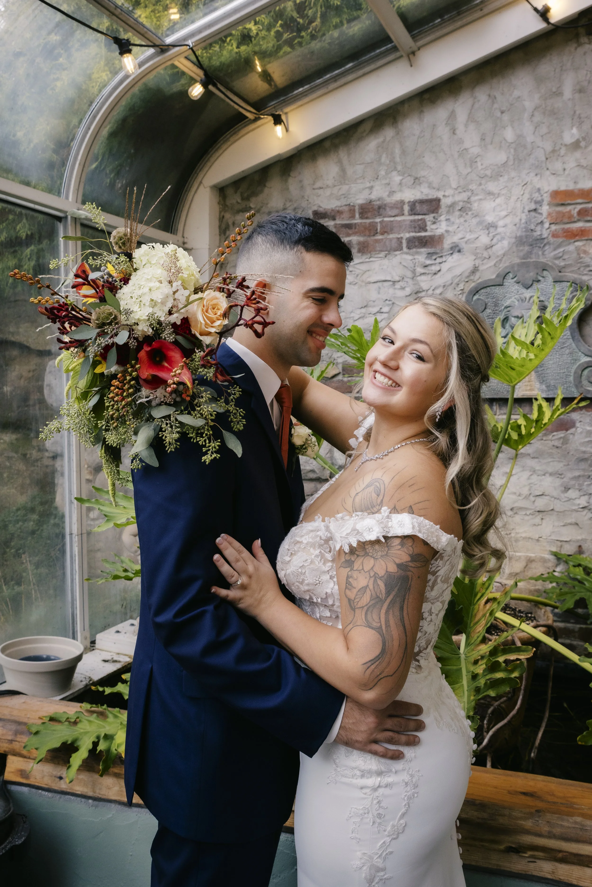 A smiling bride and groom embrace in a greenhouse with lush green plants and large windows.
