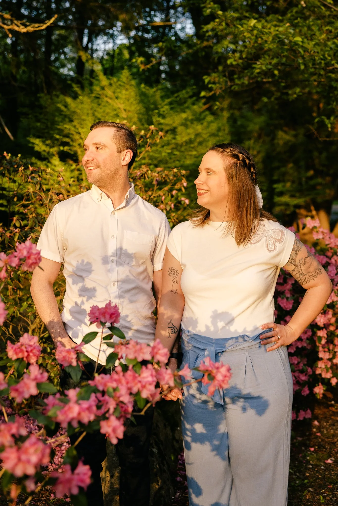 A man and woman standing outdoors in a garden with pink flowers and green foliage, holding hands and smiling at each other during sunset.