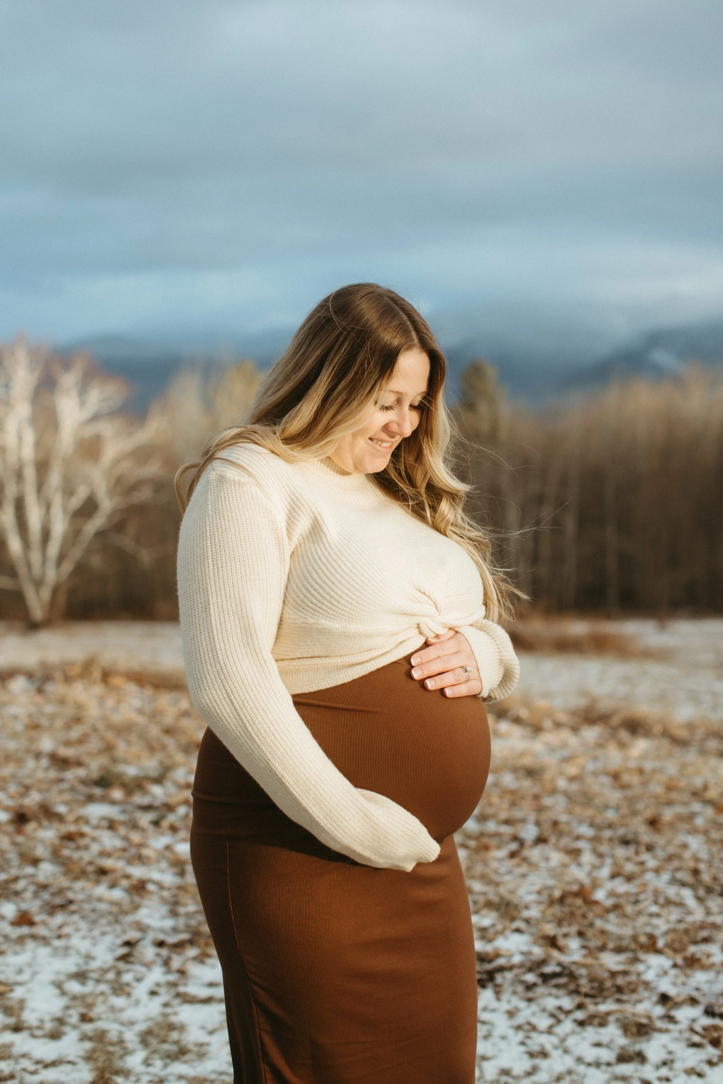 A pregnant woman with long hair smiling outdoors, holding her belly, with a background of trees, mountains, and a partly cloudy sky.