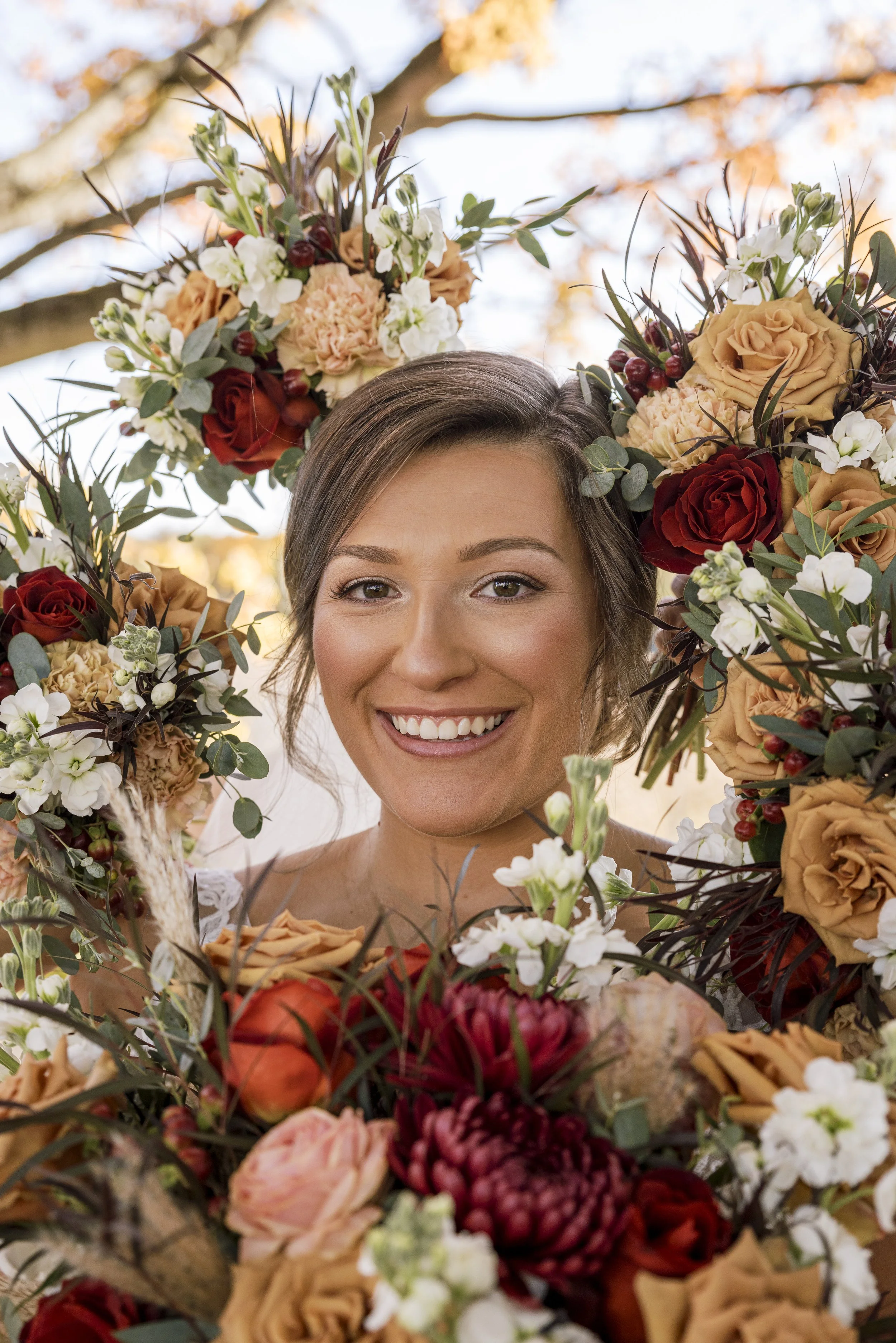 A woman smiling with a large floral wreath around her head, featuring roses, peonies, and greenery, set outdoors with a background of trees and sky.