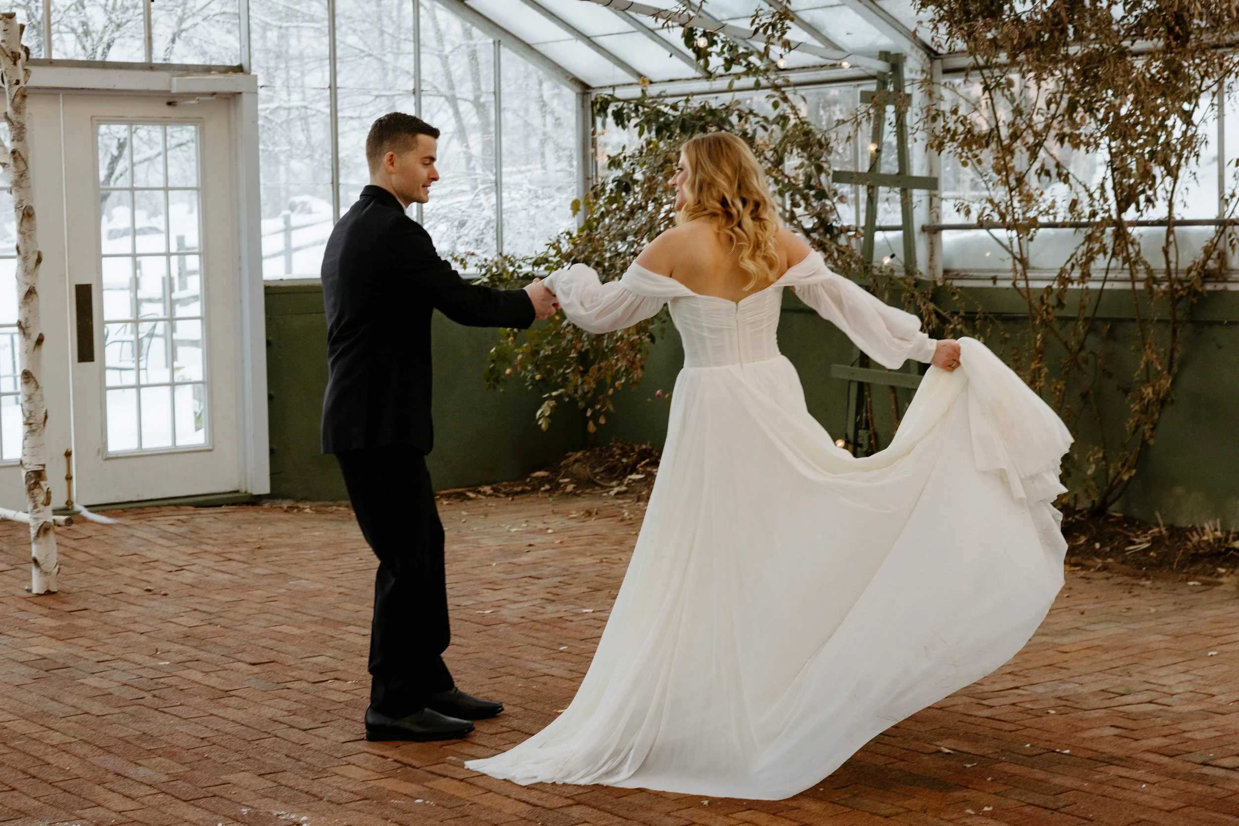 A couple, dressed in formal wedding attire, is holding hands and dancing inside a glass-enclosed space with snow visible outside. The bride is wearing a white wedding gown, and the groom is in a black suit.