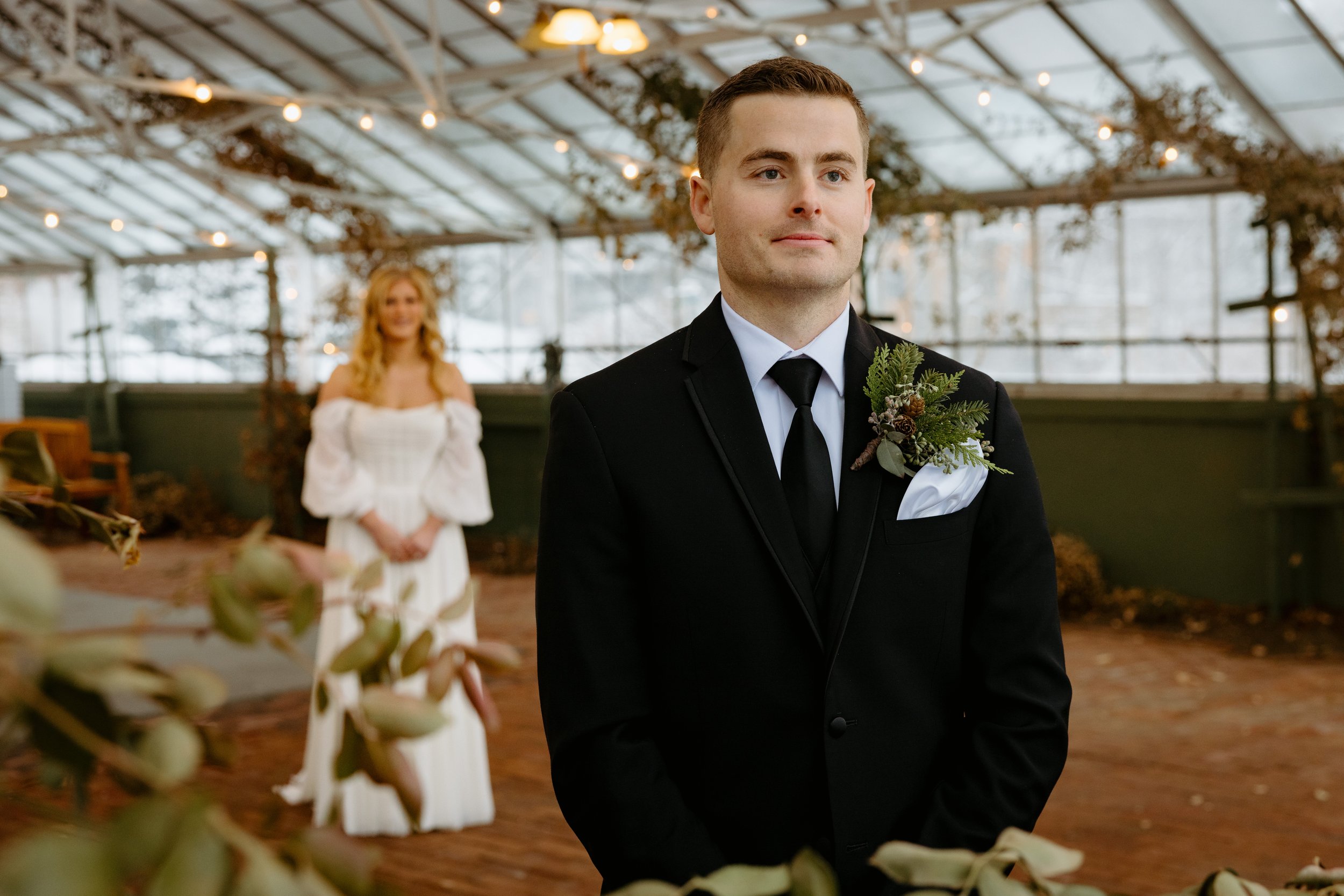 A man in a black tuxedo with a boutonniere stands in the foreground of a greenhouse-like venue, with a woman in a white dress in the background.