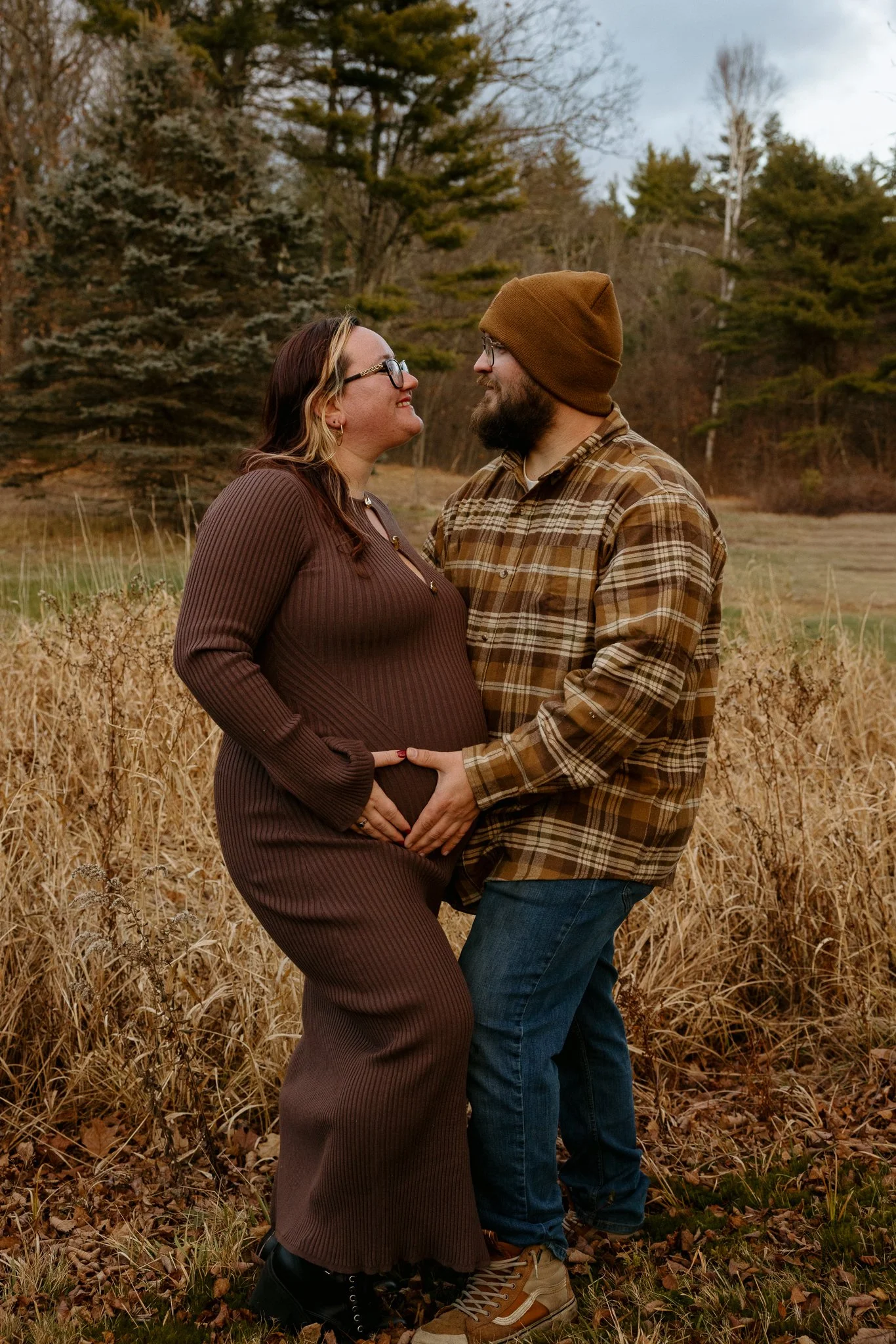 A pregnant woman and a man standing outdoors in a field surrounded by trees, holding hands and smiling at each other.