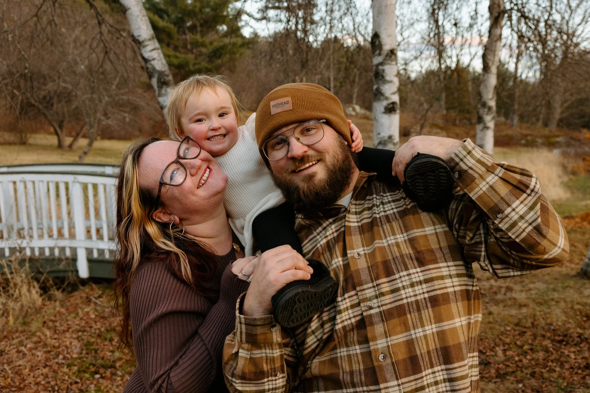 Three people outdoors in autumn, a woman with glasses, a man with glasses and beard, and a young girl smiling with her arms around their necks, in front of a white fence and trees with bare branches.