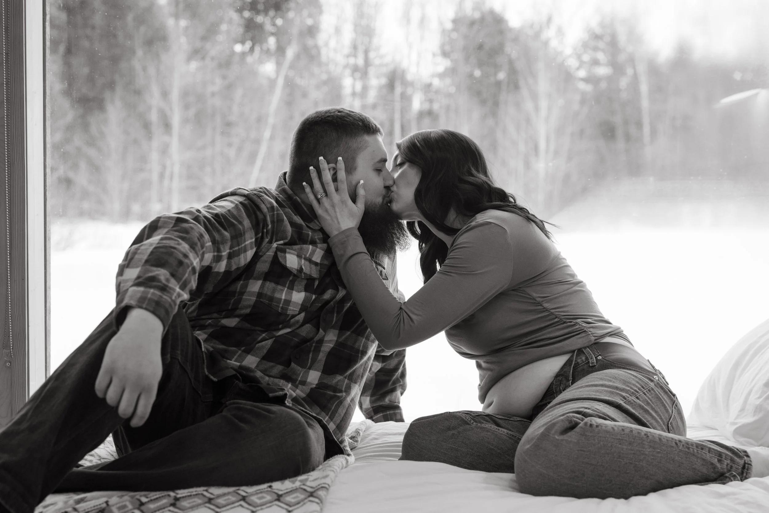 A black and white photo of a couple kissing on a bed, with the woman holding the man's face. The man has a beard and short hair, and the woman has long dark hair. Behind them is a window with trees outside.