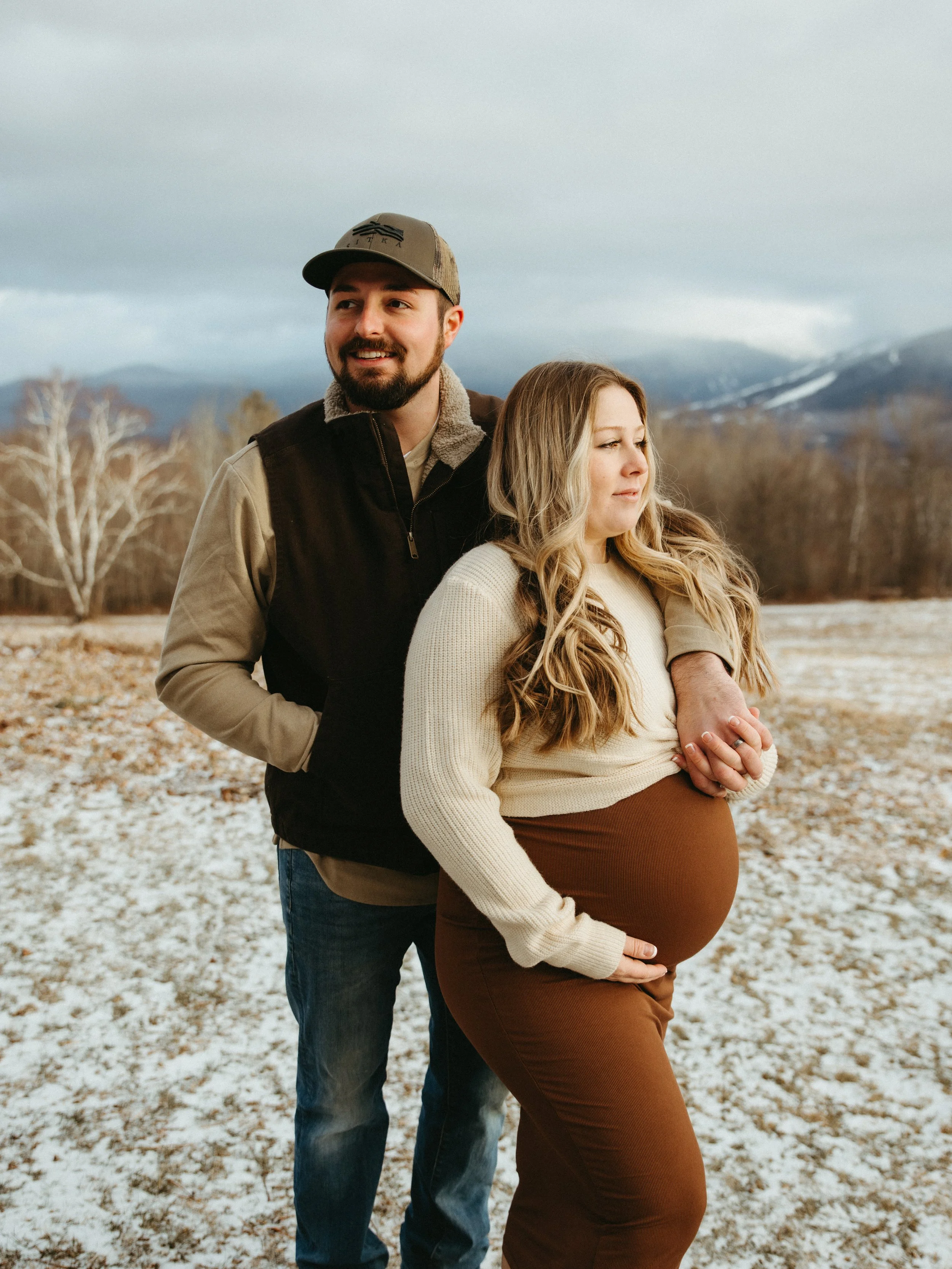A man and pregnant woman standing outdoors in a field with snow and mountains in the background. The man is smiling and has his arms around the woman, who is gently holding her belly.