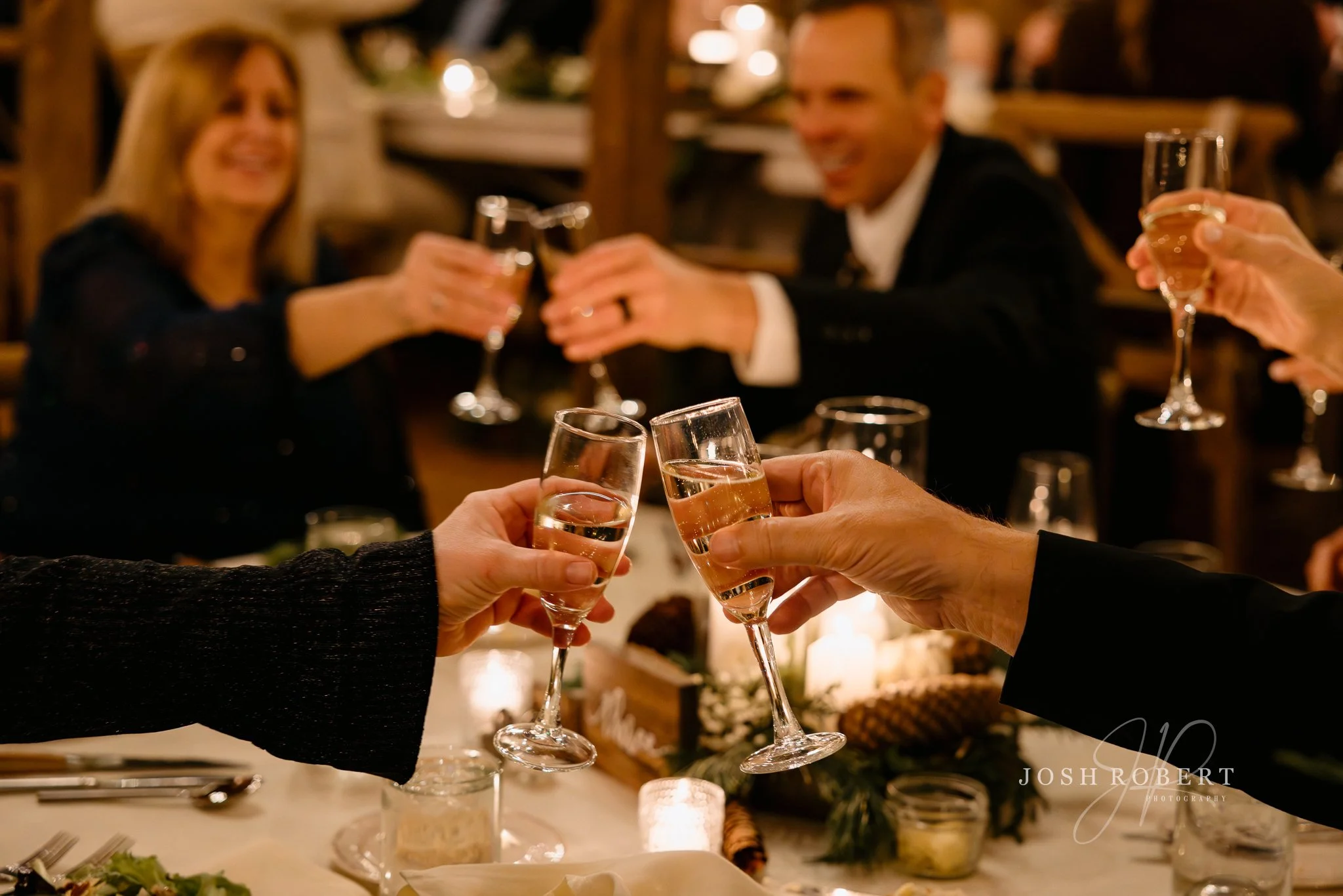 People at a dinner table raising wine glasses for a toast in a warmly lit restaurant, with a blurred background and a lit candle on the table.