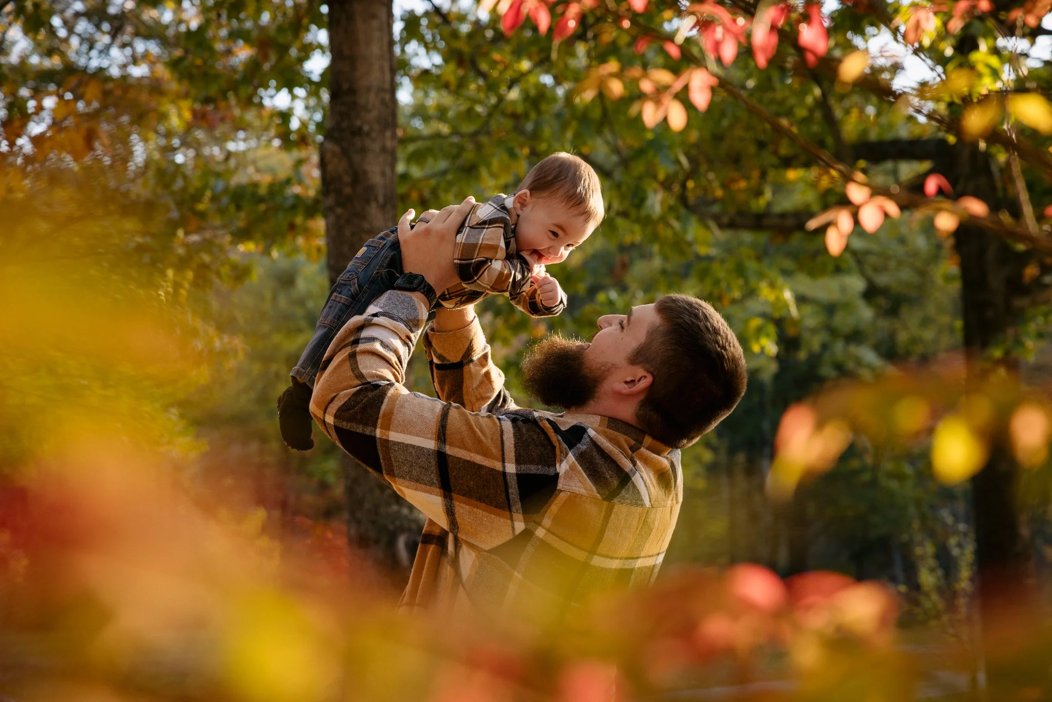 A father lifting his smiling young son in a park during autumn, surrounded by colorful fall leaves.