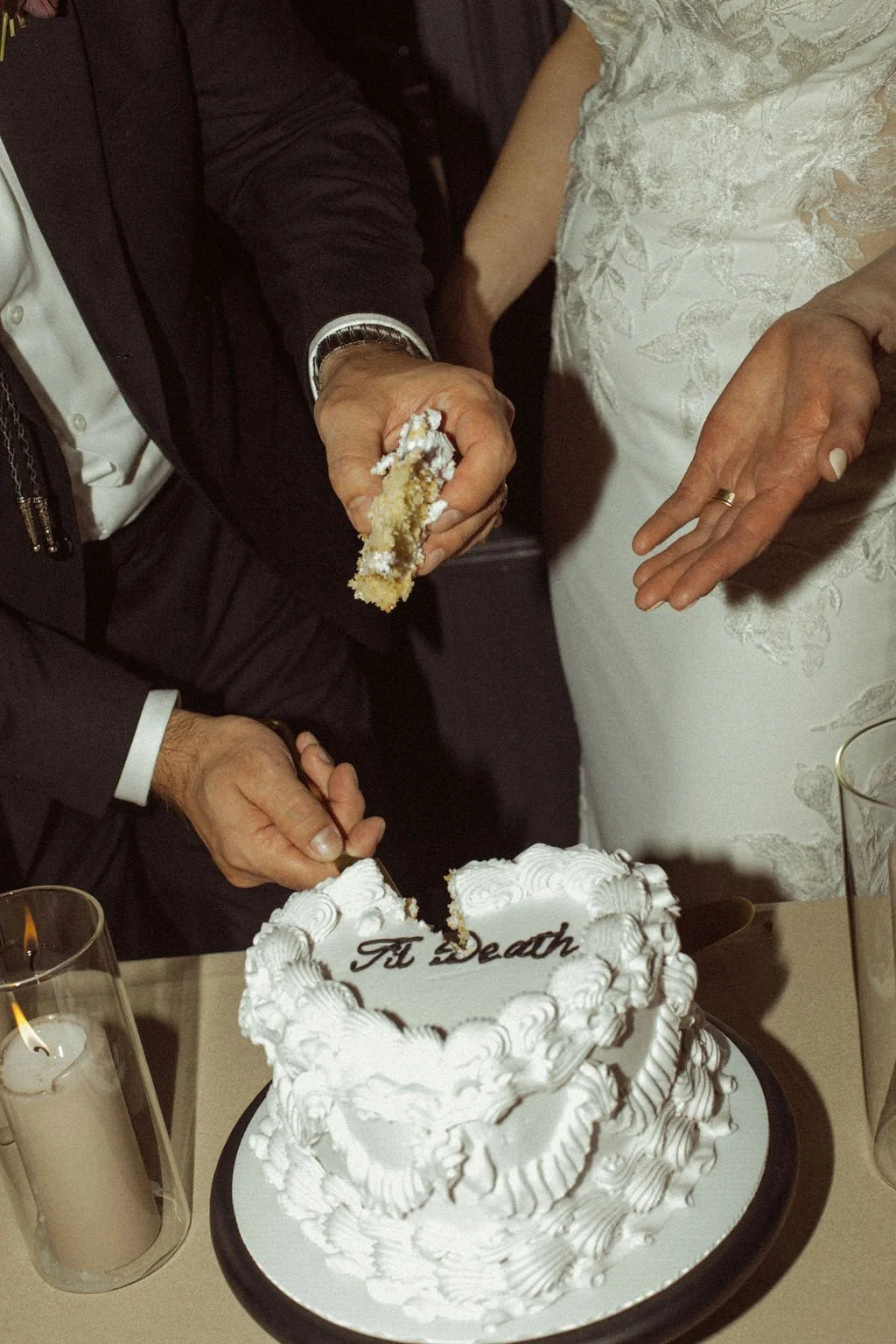 A wedding cake with the words 'Till Death' written on it, and two people cutting a piece of the cake together at a wedding celebration. Brides. Grooms. Bride and Groom. Couples. Weddings. Lifestyle. Family. Friends. Festivals. Wedding Photographer. 