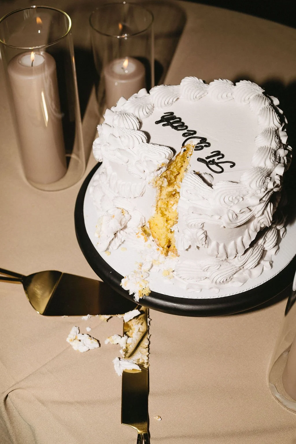 A partially sliced white frosted birthday cake with black writing on top, placed on a black cake stand, with cake crumbs and a part of the slice on a cake server. In the background, three lit candles in glass holders are visible.
