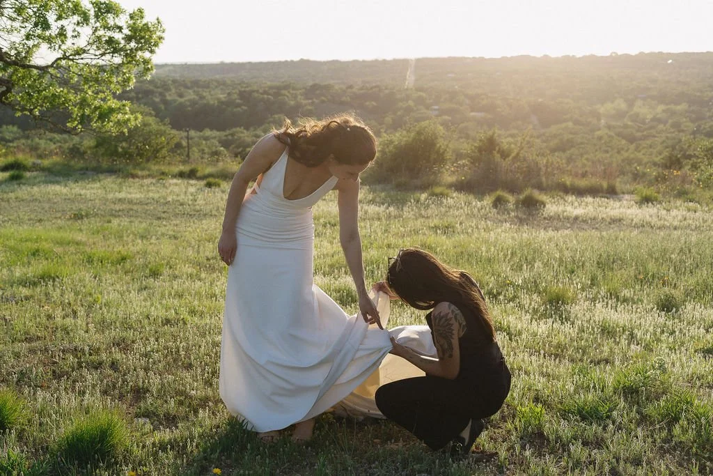 A woman in a white dress with long hair bent over a woman in dark clothing with long hair, who is sitting and holding a piece of fabric in a grassy field during sunset. LGBTQIA photographer.