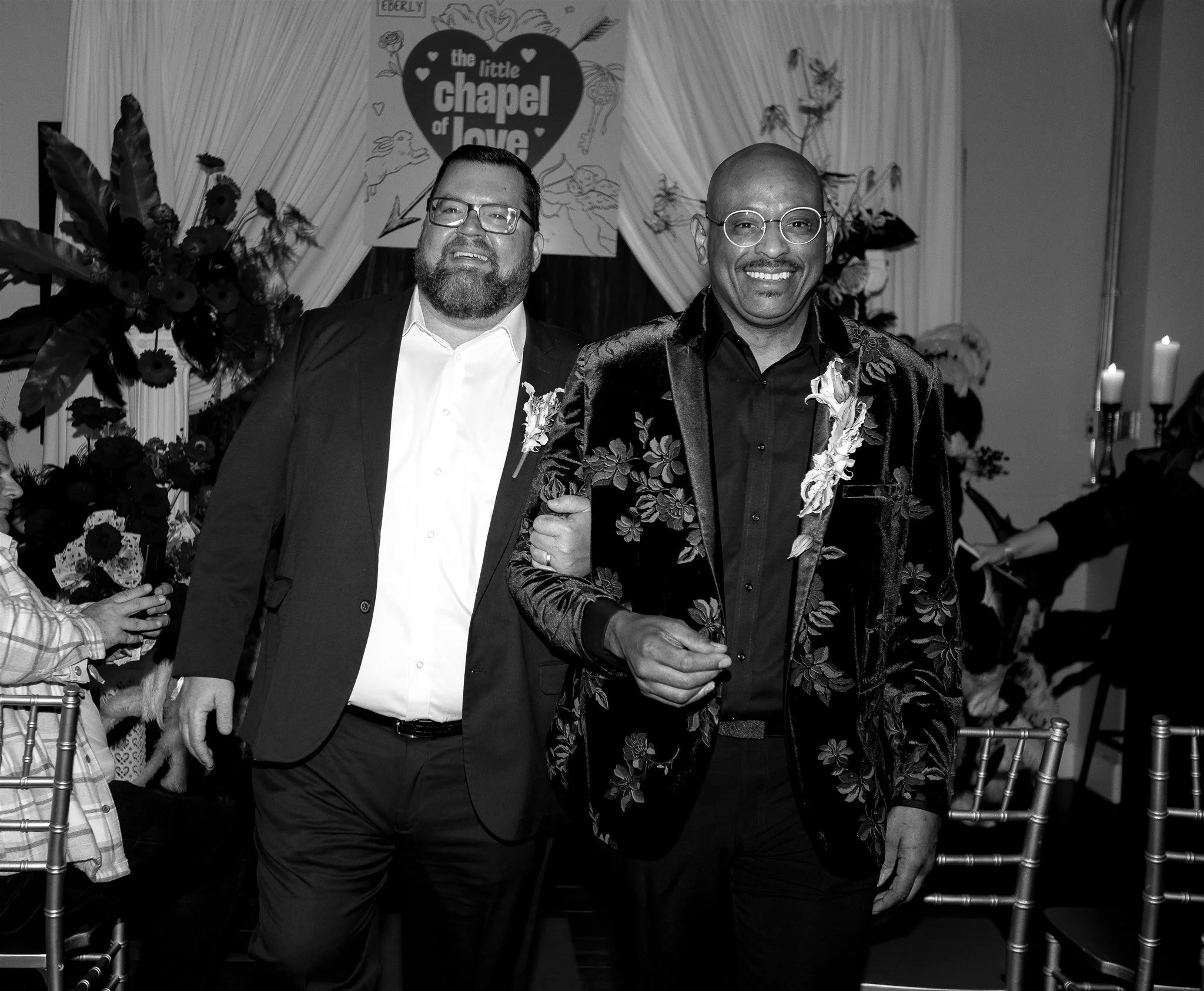 Two men dressed in wedding attire, smiling as they walk together. The background features floral decorations, candles, and a sign that says, 'the little chapel of love.' Vegas Wedding. Austin photographer. Wedding photographer. Travel photographer