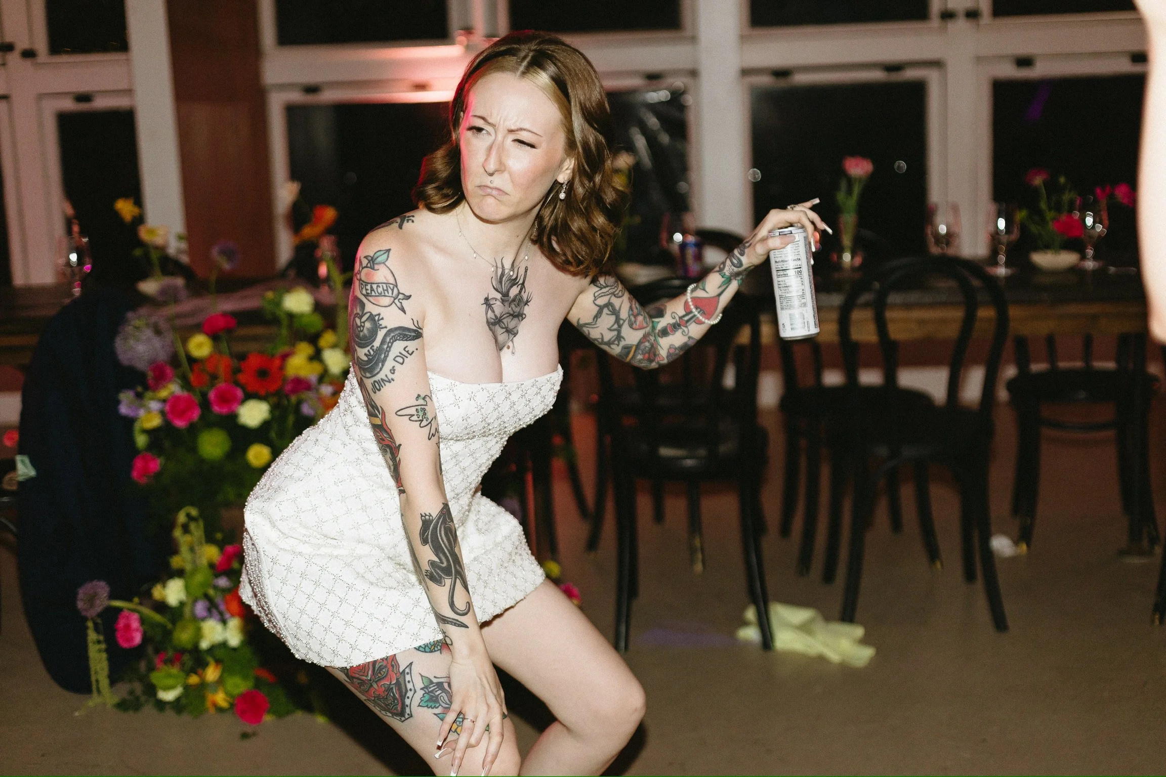 A woman with tattoos on her arms and legs wearing a white strapless dress, holding a can of spray, in a room with flowers, chairs, and a large window.
