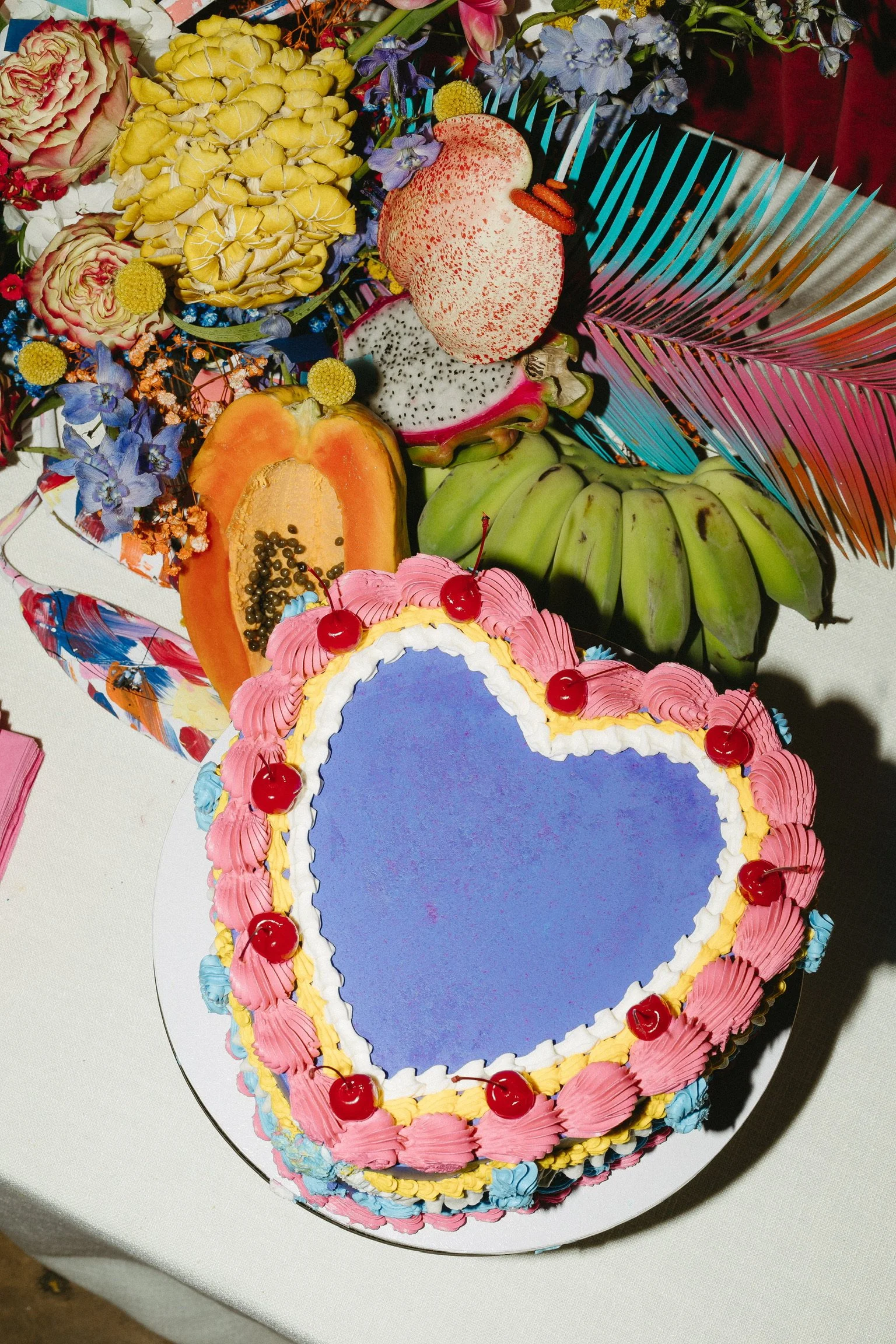 Heart-shaped birthday cake with pink and blue frosting and cherries on top, on a table with various fruits and colorful flowers.