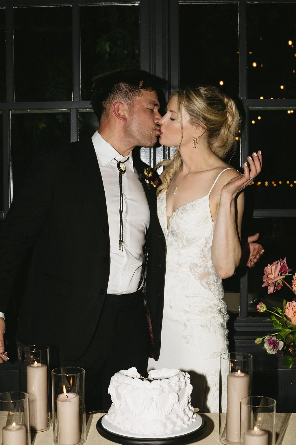 A newlywed couple shares a kiss at their wedding reception, standing behind a wedding cake with lit candles in glass holders and floral decorations in the background.