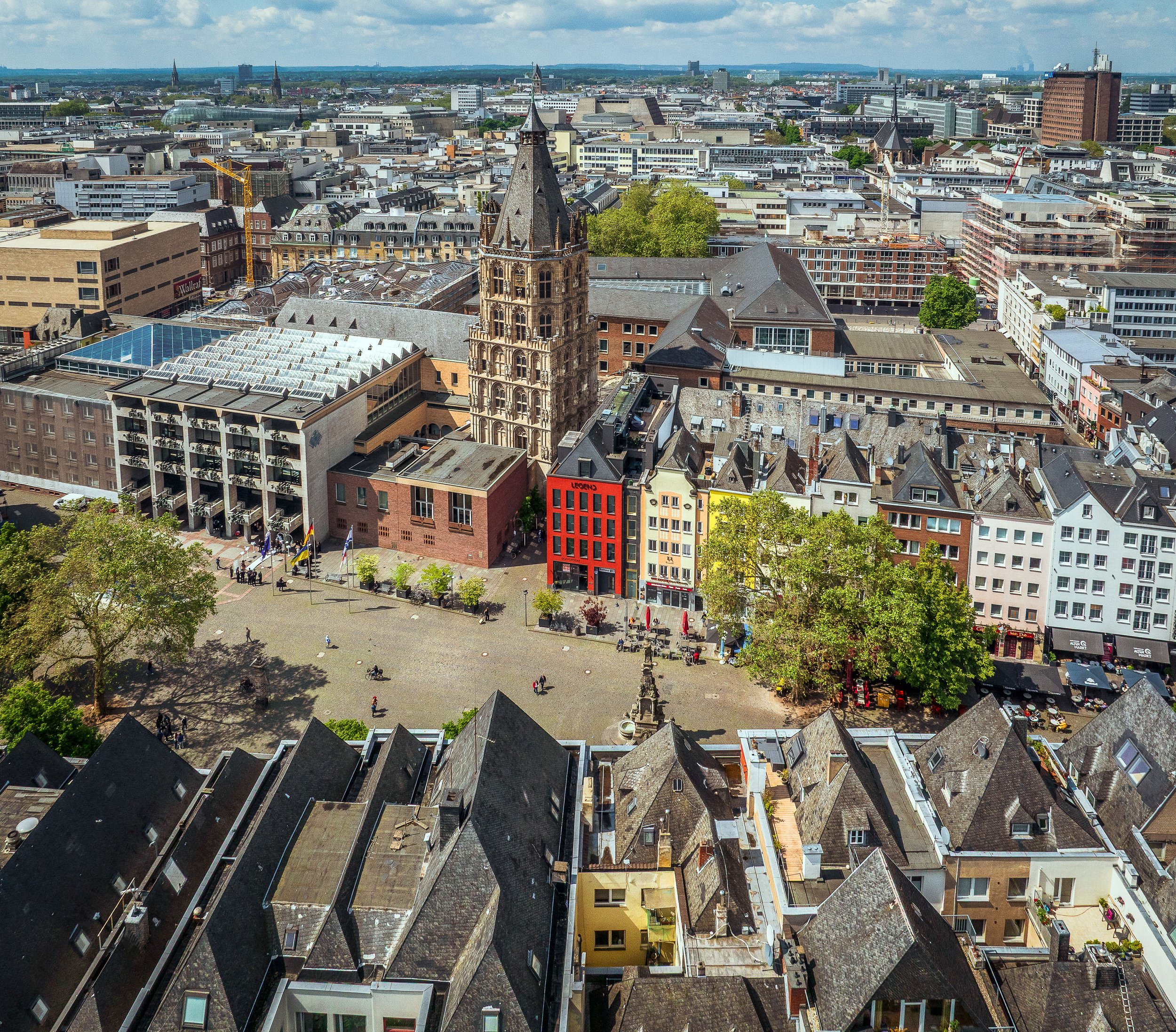 Luftaufnahme des Alter Markts mit dem historischen Rathausturm, bunten Altstadthäusern und dem Jan-von-Werth-Brunnen. Rund um den weitläufigen Platz sind Menschen, Außengastronomie und die dichte Bebauung der Kölner Innenstadt zu sehen.