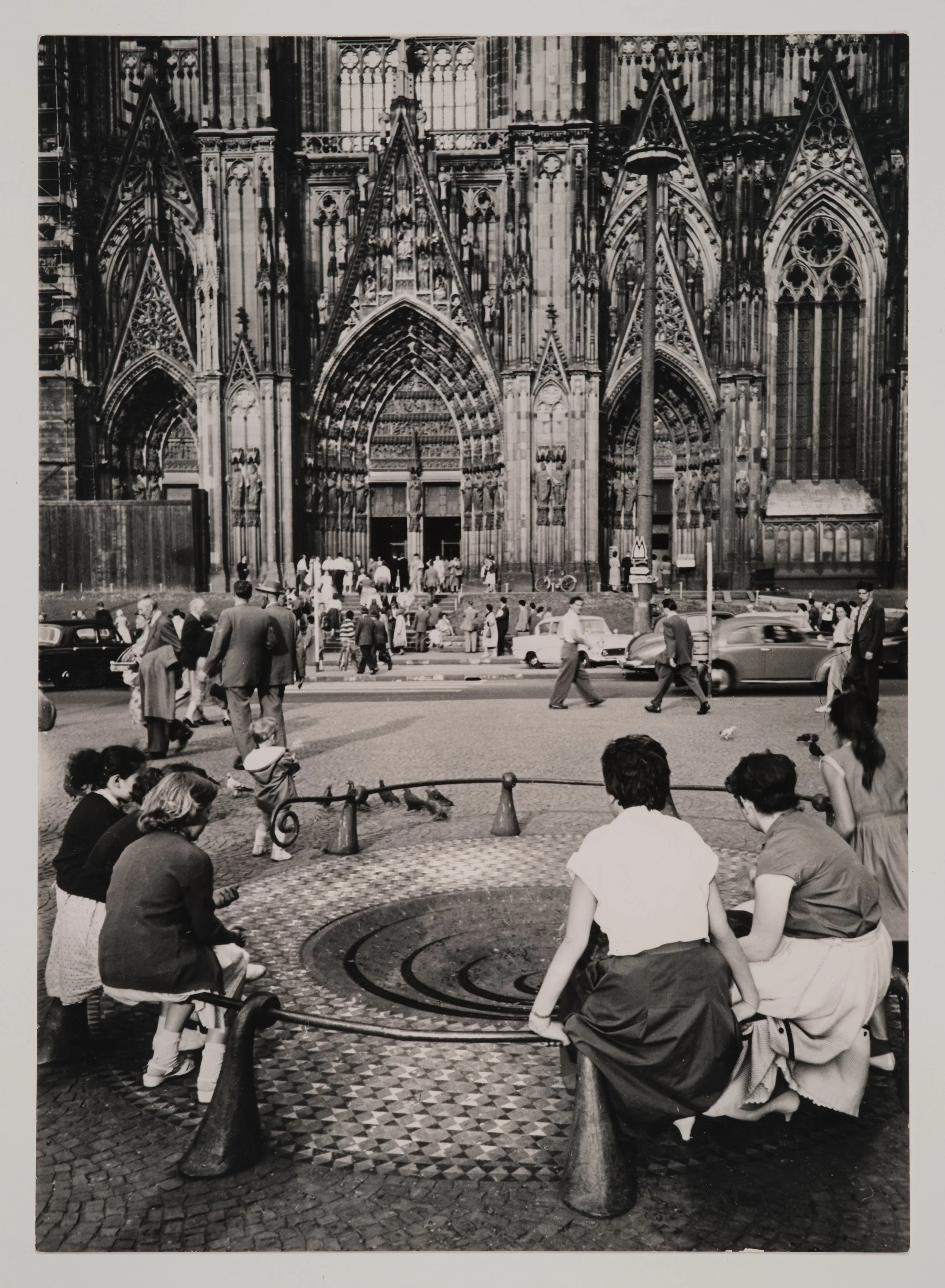 Historische Schwarz-Weiß-Aufnahme des Taubenbrunnens auf der Kölner Domplatte. Menschen sitzen am Brunnen und blicken auf den Kölner Dom im Hintergrund, während Passant*innen und Autos den Platz beleben.