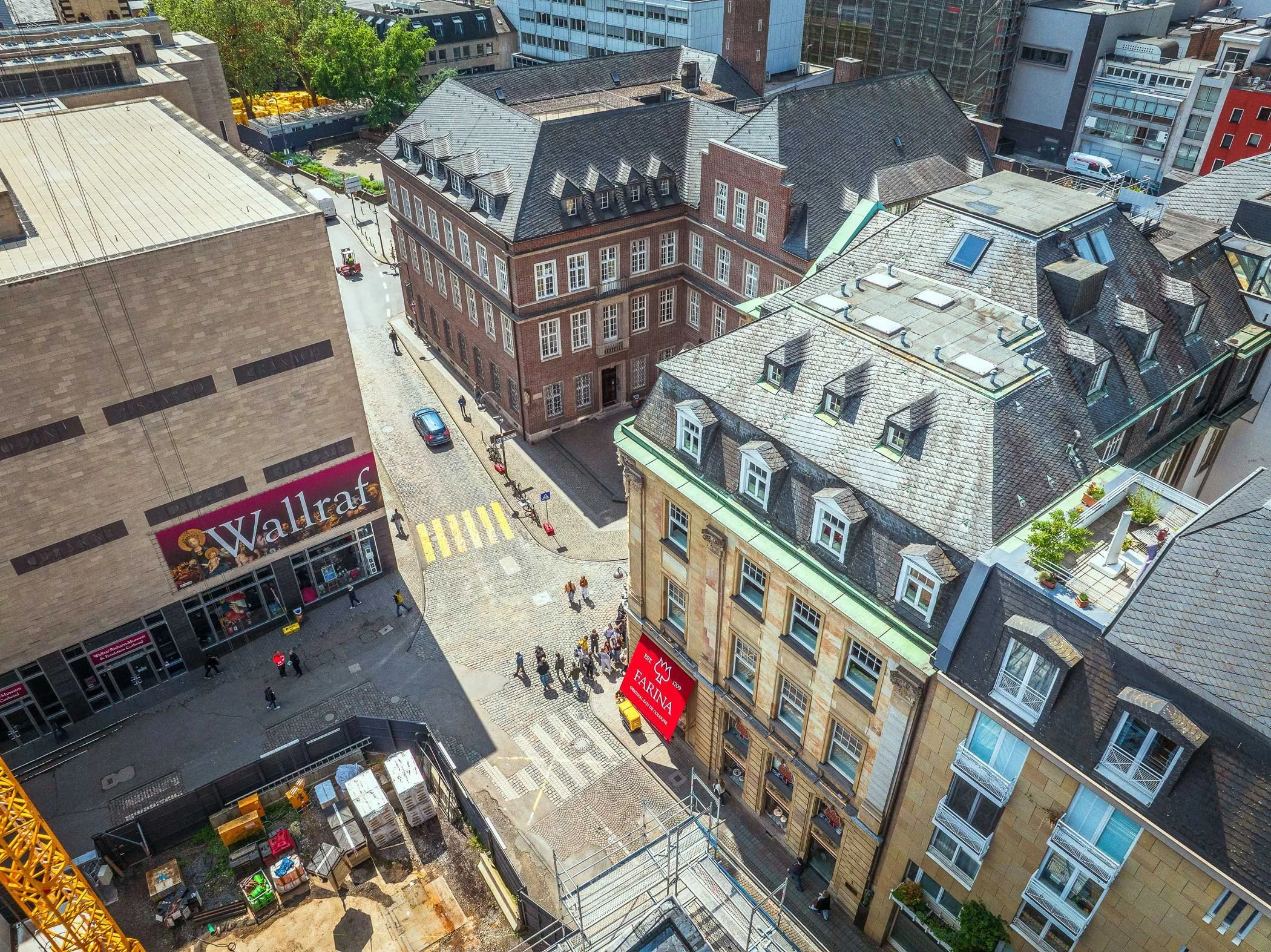 Luftaufnahme des Gülichplatzes in der Kölner Altstadt mit dem Wallraf-Richartz-Museum links, dem Farina-Haus rechts und historischen Gebäuden entlang der gepflasterten Straße. Im unteren Bildbereich ist eine Baustelle zu sehen, mehrere Menschen überq