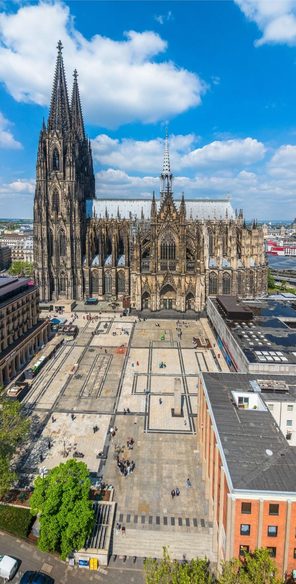 Luftaufnahme des Roncalliplatzes vor dem Kölner Dom bei klarem Himmel. Die beiden Domtürme prägen das Bild, während sich der weitläufige Platz zwischen Dom, Museum Ludwig und den umliegenden Gebäuden erstreckt.