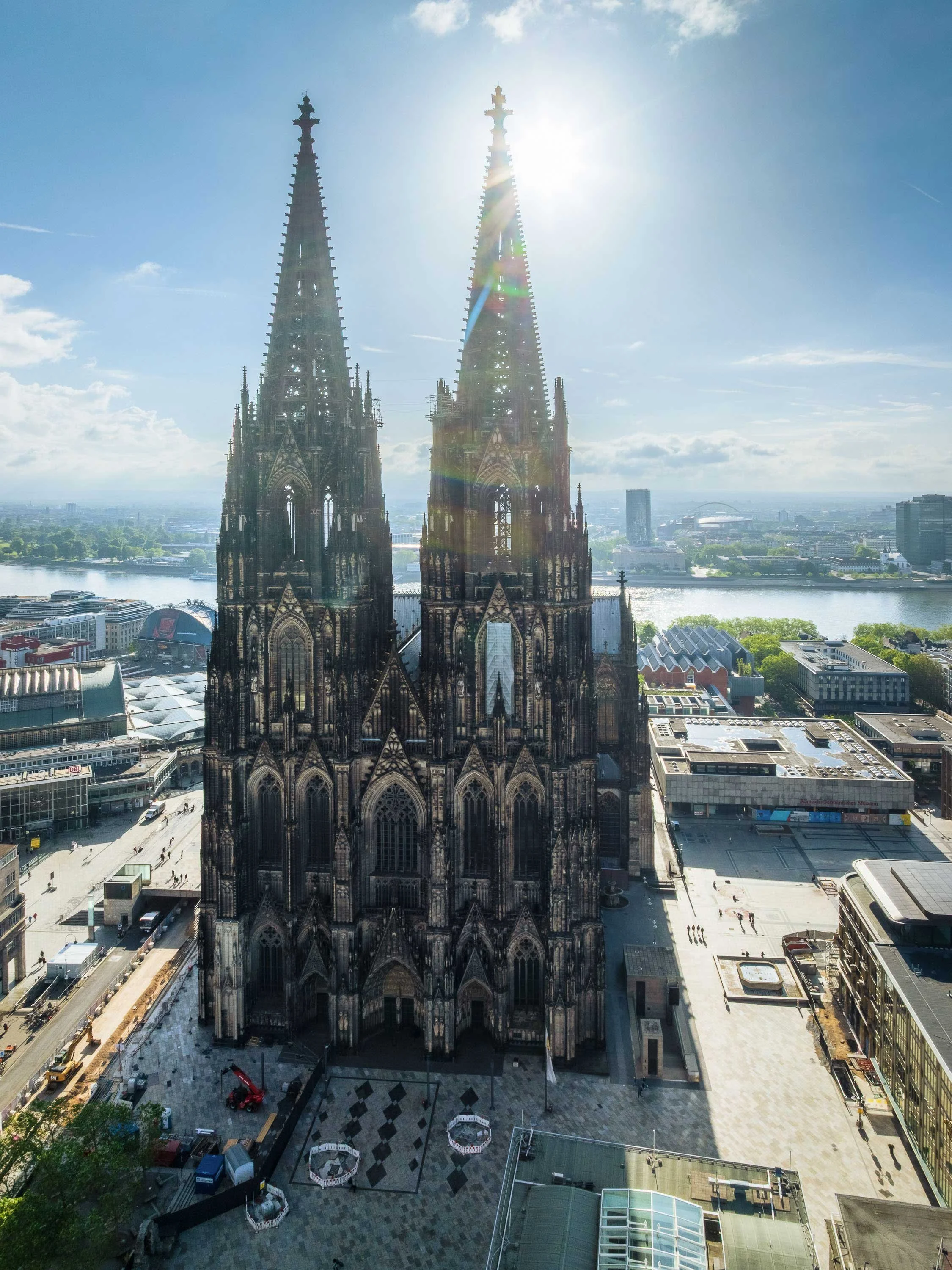 Frontale Luftaufnahme des Kölner Doms am Domkloster bei strahlendem Sonnenschein. Die beiden gotischen Türme ragen vor blauem Himmel auf; dahinter sind der Rhein, die Hohenzollernbrücke und Teile der rechten Rheinseite zu sehen.
