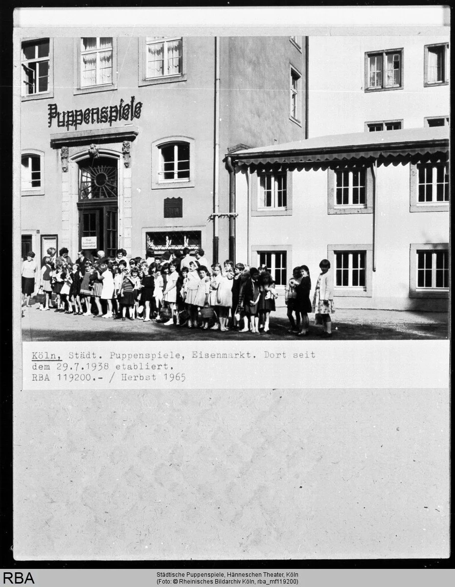 Historische Schwarz-Weiß-Fotografie des Hänneschen-Theaters am Eisenmarkt in Köln (1965). Vor dem Eingang mit der Aufschrift „Puppenspiele“ warten zahlreiche Kinder und Erwachsene auf den Einlass.